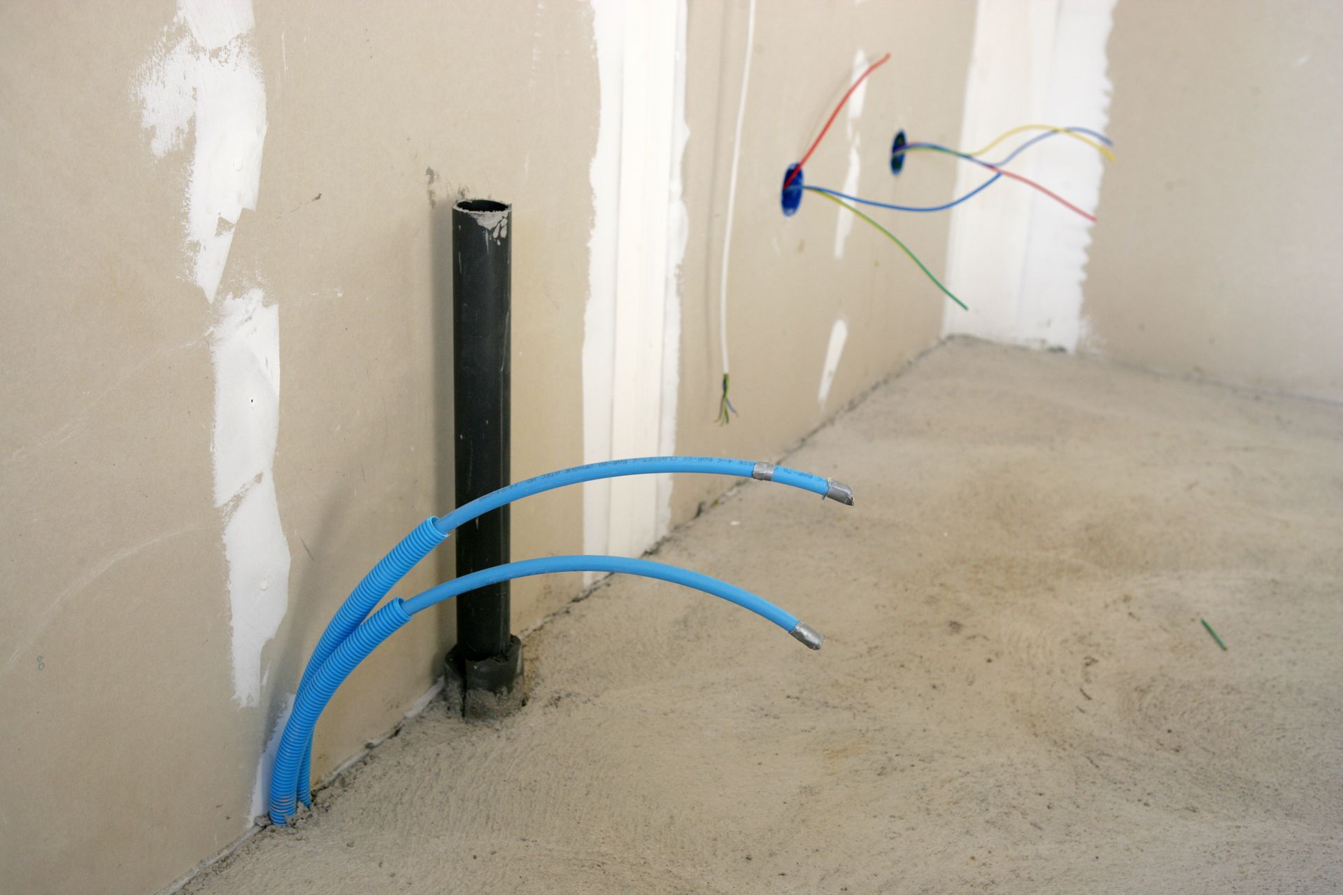 Chantier de construction avec tuyauterie et câblage électrique apparents dans un coin d'une pièce aux murs en plaques de plâtre.