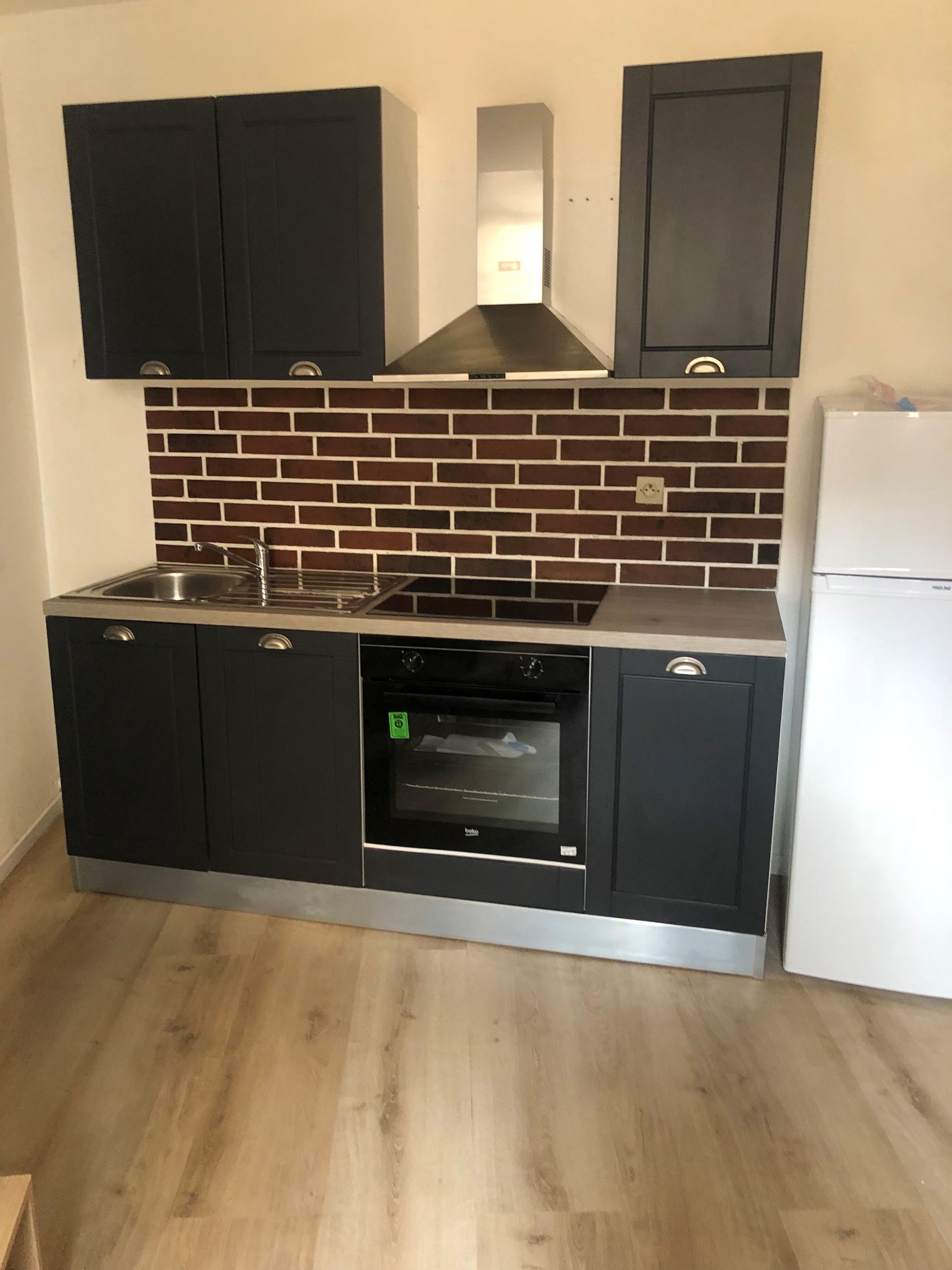 Petite cuisine avec placards foncés, crédence en briques, hotte en inox et réfrigérateur.