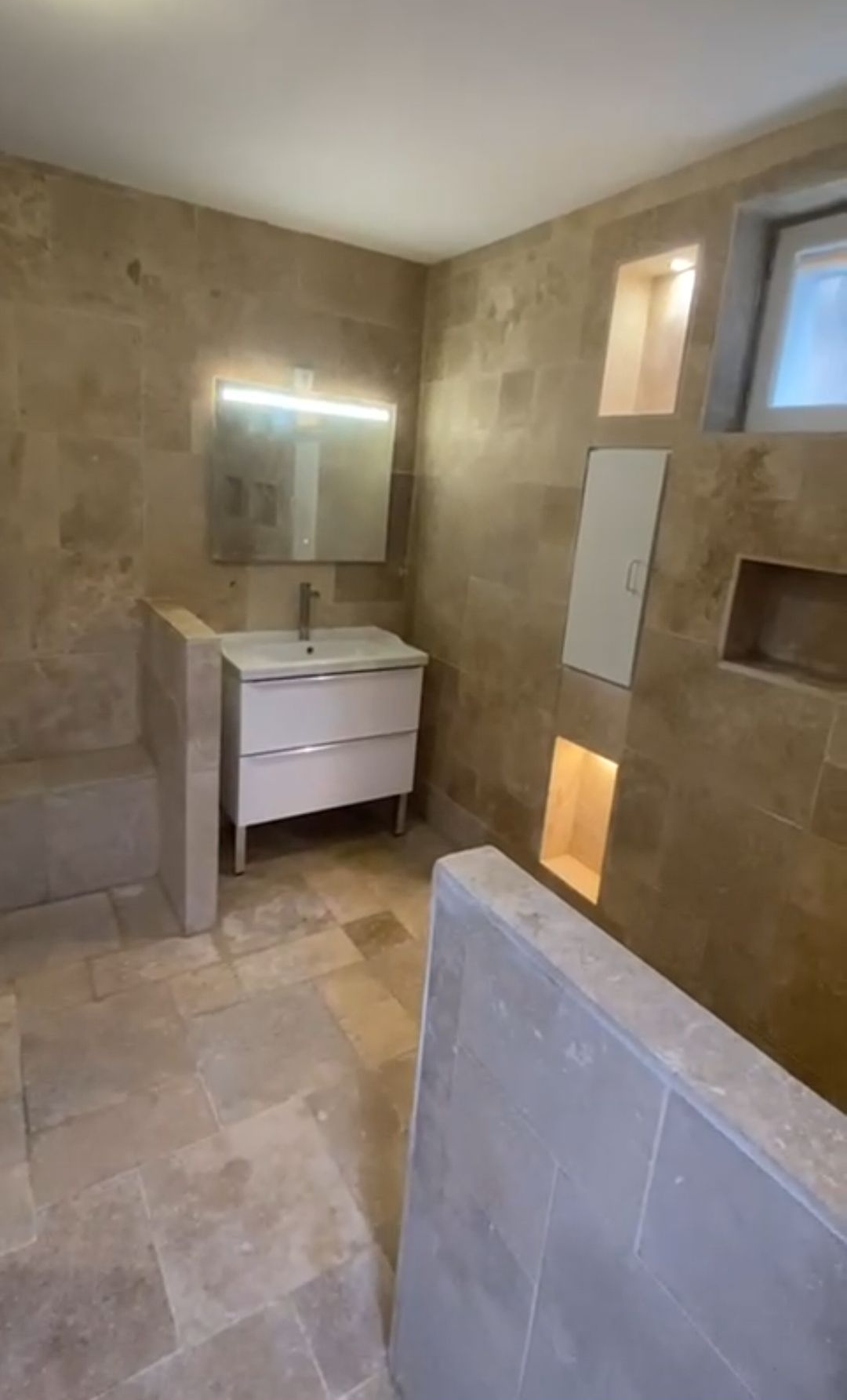 Salle de bains avec murs et sol carrelés de beige. Meuble-lavabo blanc, miroir et étagères intégrées.