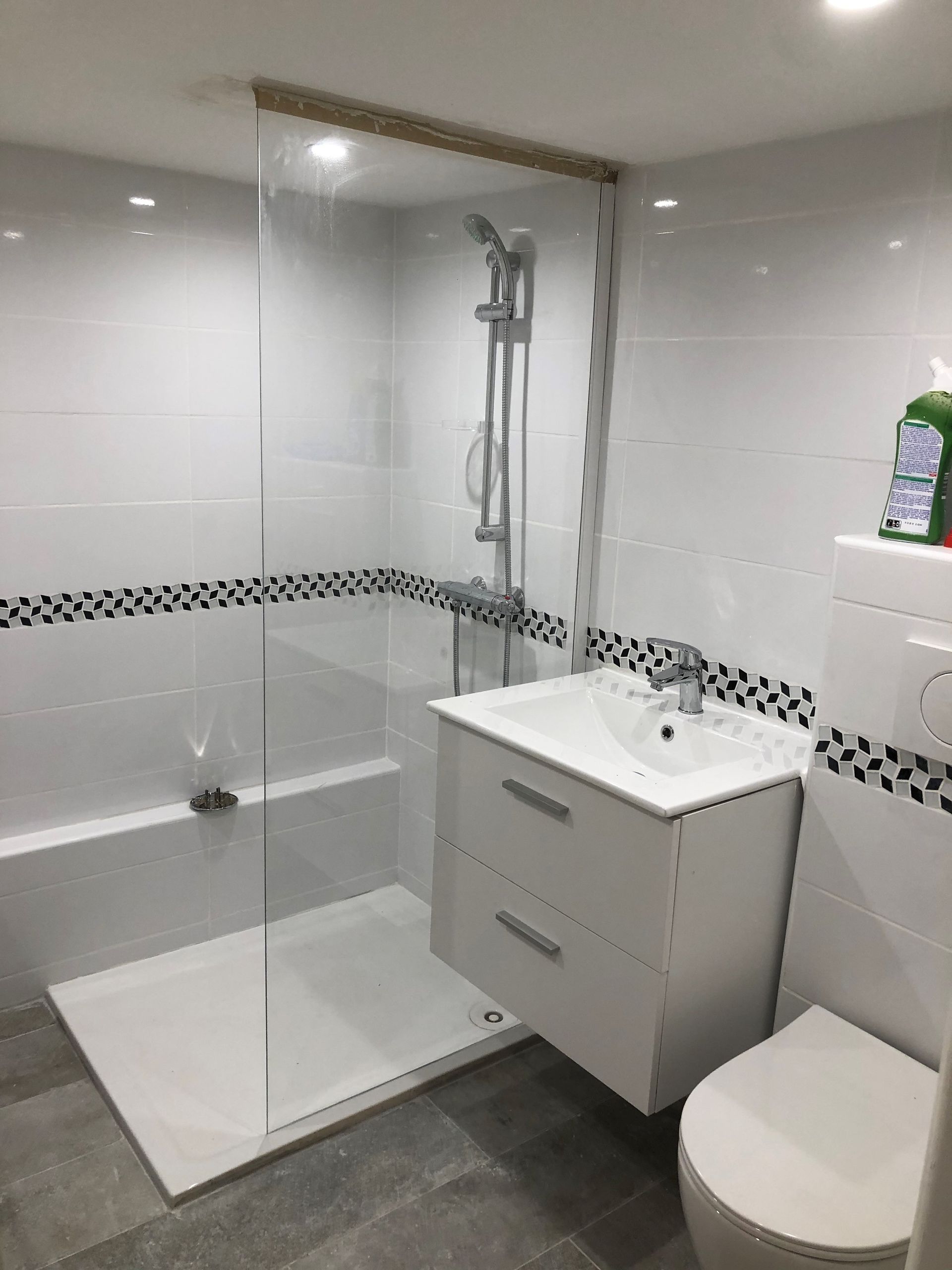 Salle de bains moderne, blanche avec douche et meuble-lavabo. Sol carrelé gris, accents de carrelage noir.