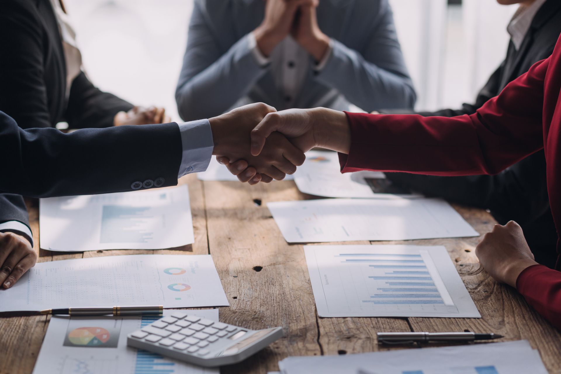 Dos personas se dan la mano sobre una mesa con papeles, calculadora y bolígrafos, rodeadas de compañeros.