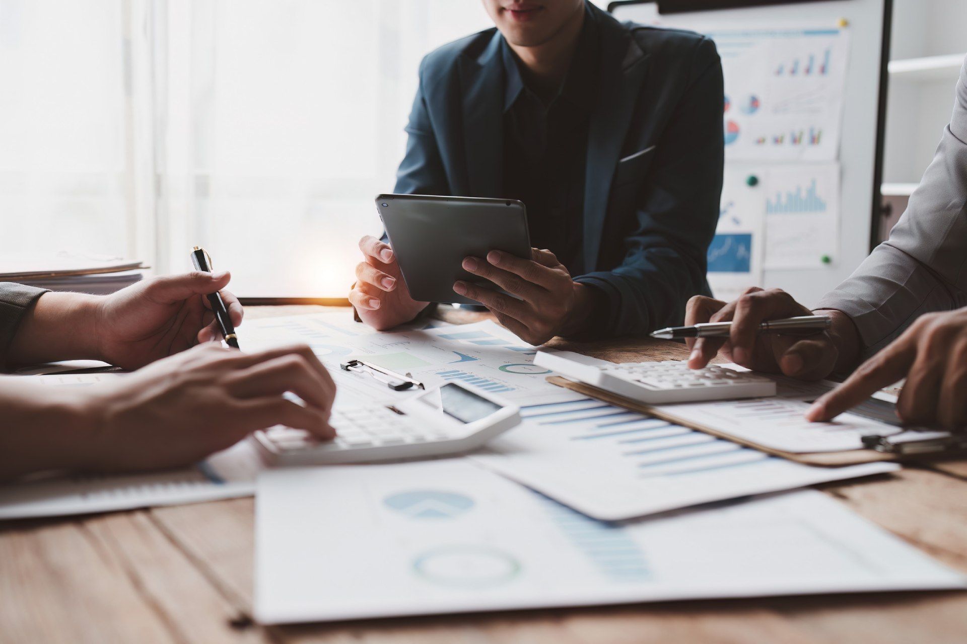 Equipo de negocios en una reunión, un hombre presentando datos en la pantalla, otros sentados a la mesa con una computadora portátil, entorno de oficina luminoso.