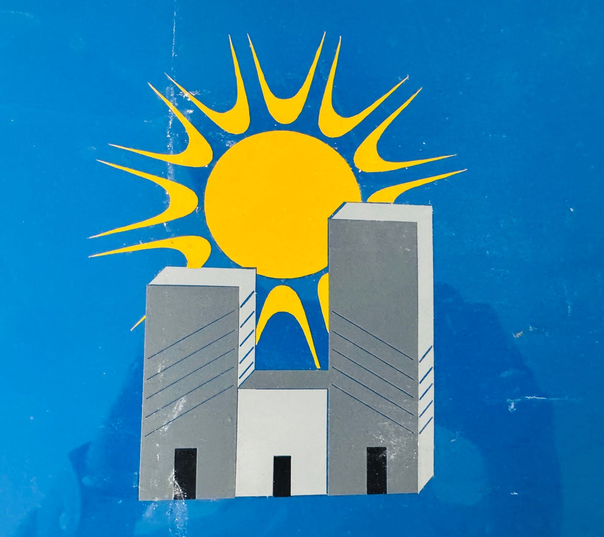 Bandera azul con un sol amarillo y dos edificios grises.