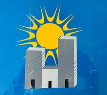 Bandera azul con un sol amarillo y dos edificios grises.