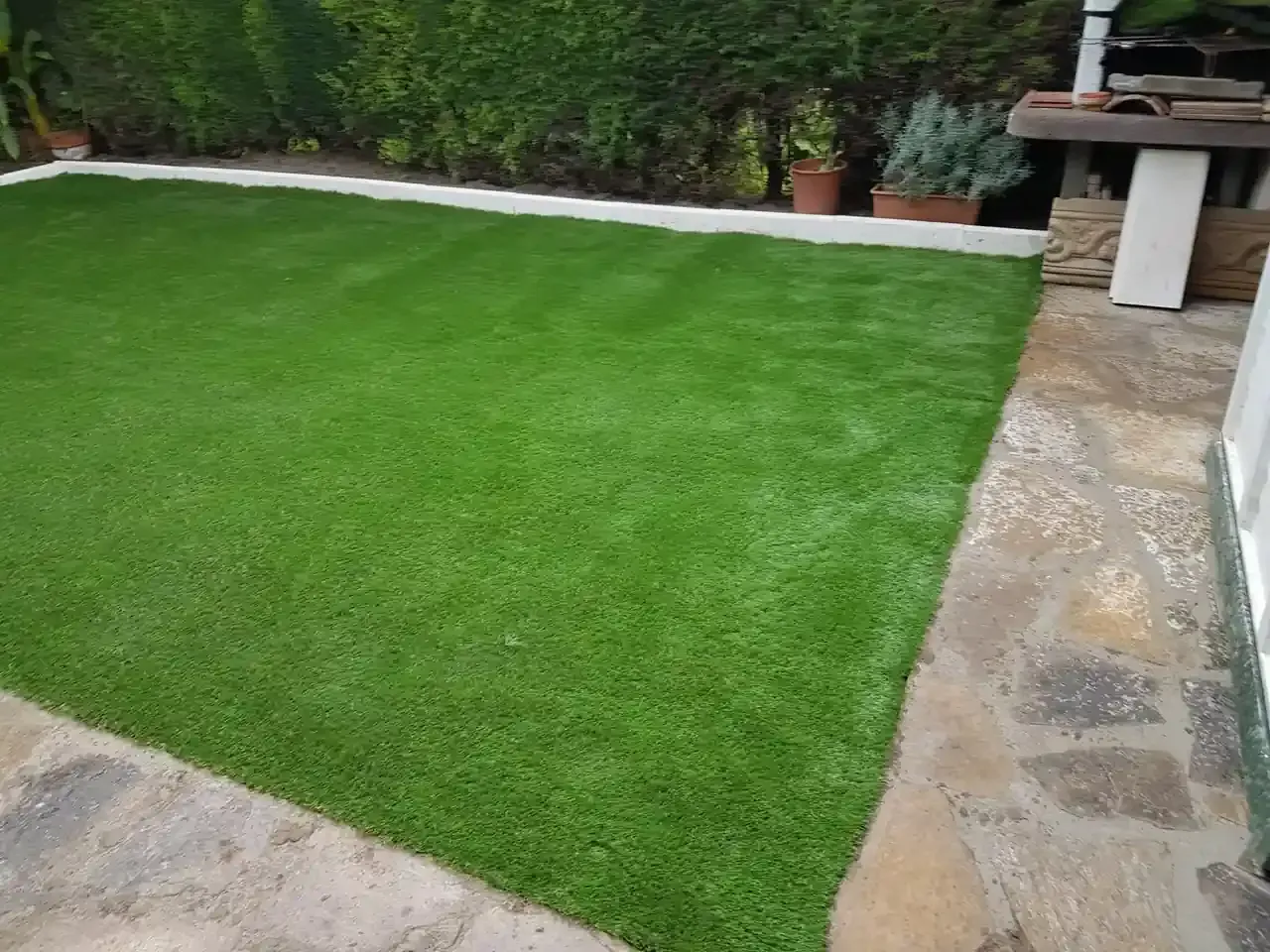 Césped artificial verde bordeado de piedra y hormigón, con arbustos al fondo.