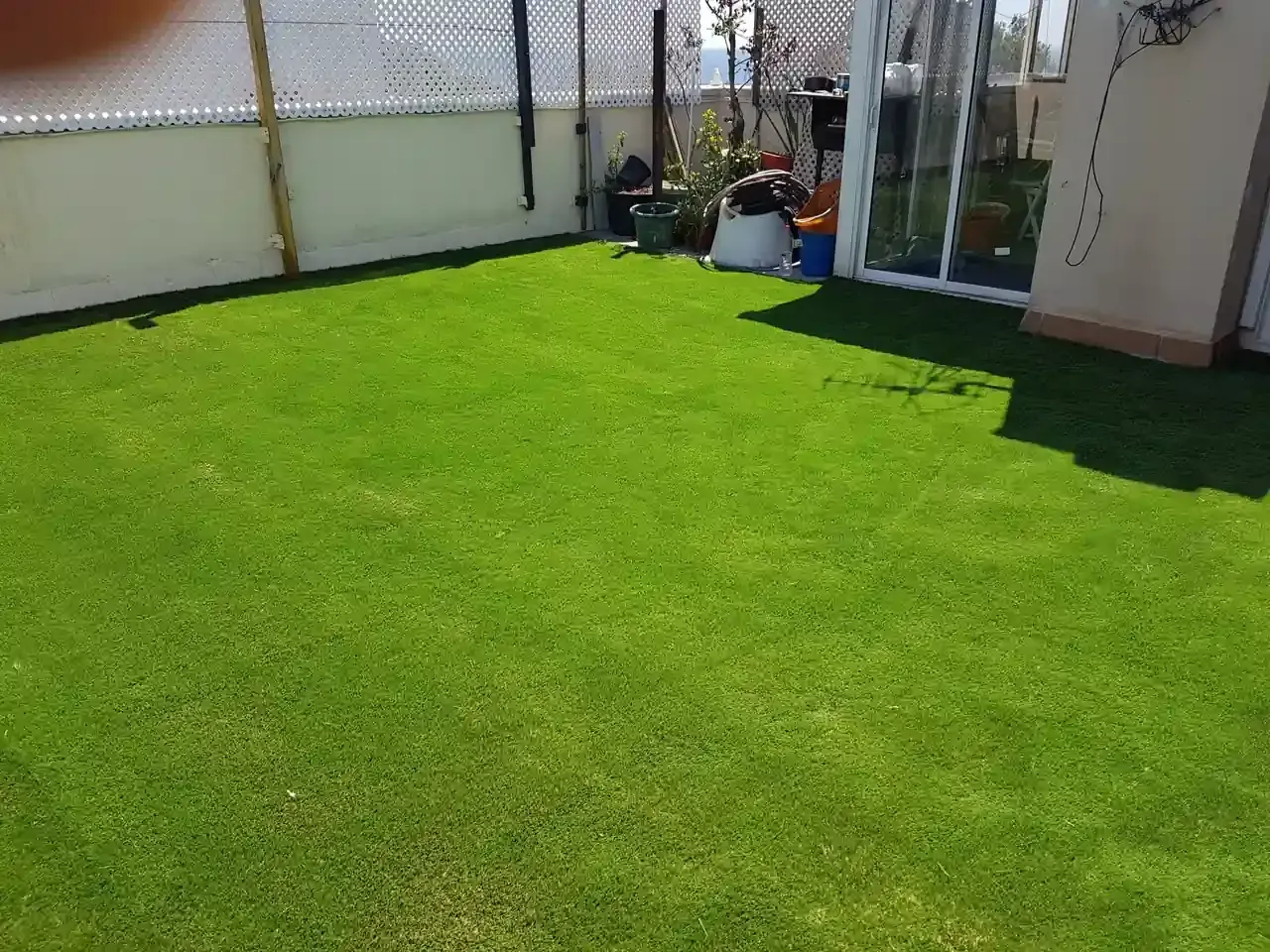 Césped artificial verde en un patio trasero al lado de una casa con una puerta de vidrio, una cerca y plantas en macetas.