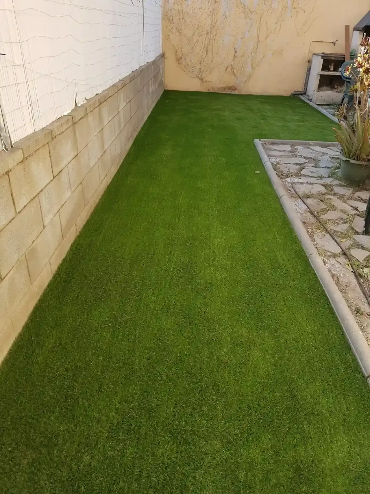 Césped artificial verde en un patio trasero estrecho junto a una pared de bloques de hormigón y un patio de piedra.
