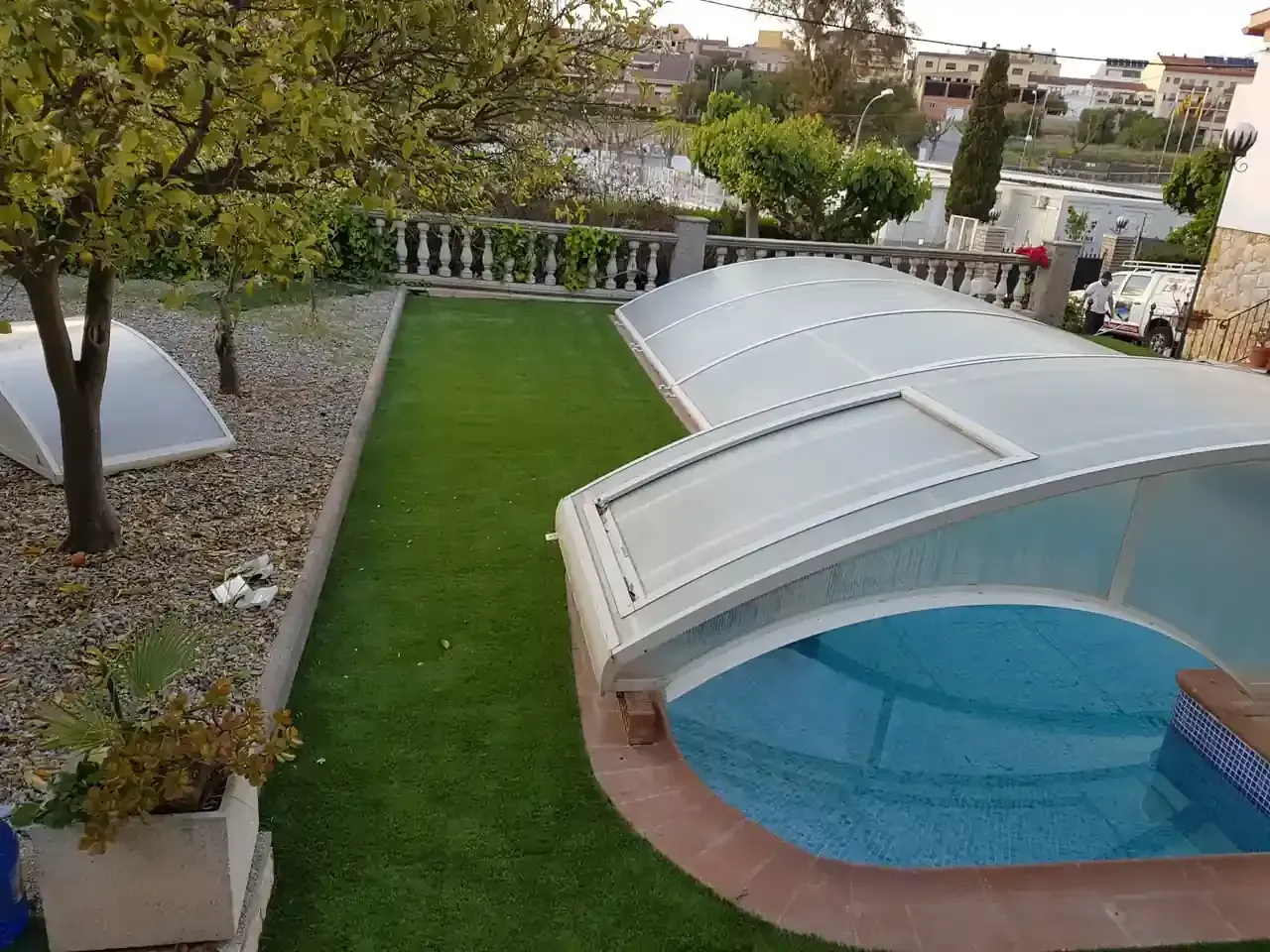 Piscina con cerramiento retráctil, césped verde, árboles y pequeña zona ajardinada.