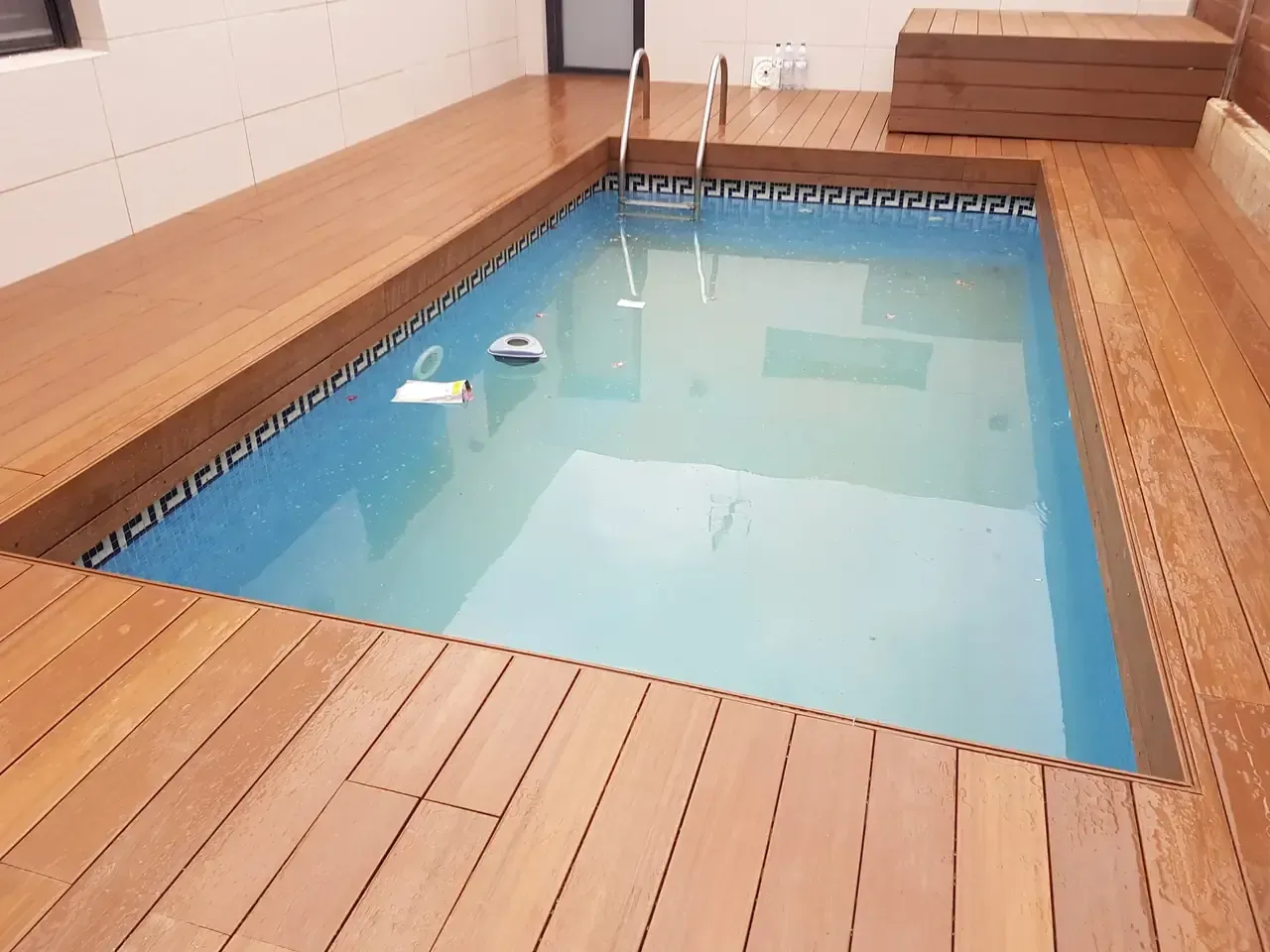 Piscina cubierta rectangular con tarima de madera. Agua azul, borde de azulejos decorativos y escalera de acero inoxidable.