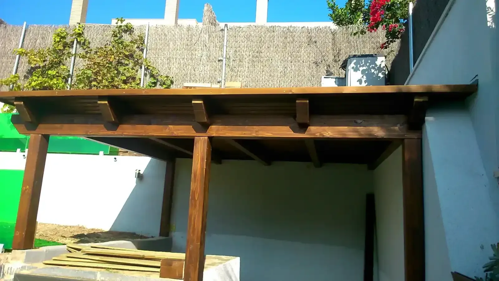 Pérgola de madera con tinte marrón, construida contra una pared blanca en un entorno exterior soleado.