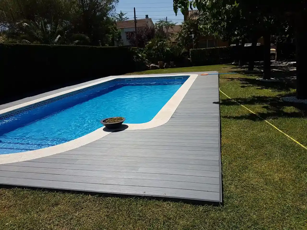 Piscina rectangular con tarima gris en un patio con césped bajo un cielo soleado.