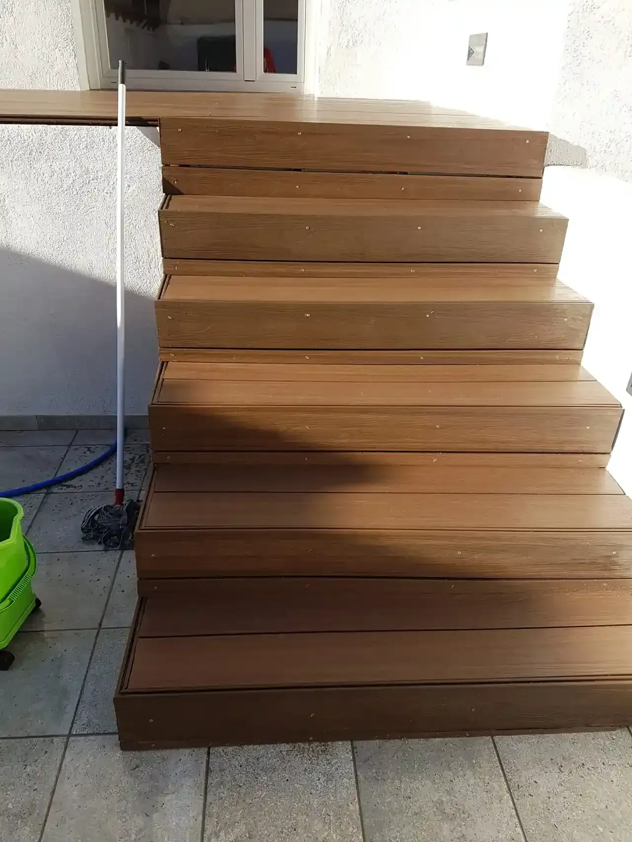 Escaleras exteriores de madera marrón que conducen a una pared blanca y una ventana. Hay un trapeador apoyado en los escalones.