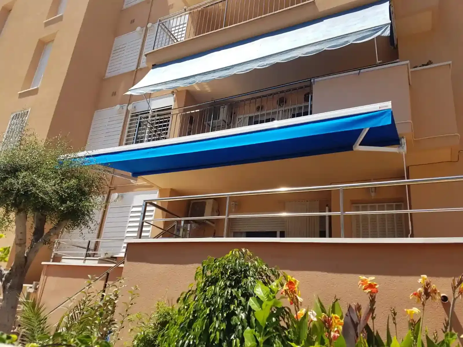 Un edificio de varias plantas con toldos azules y blancos en los balcones. Exterior soleado con vegetación.