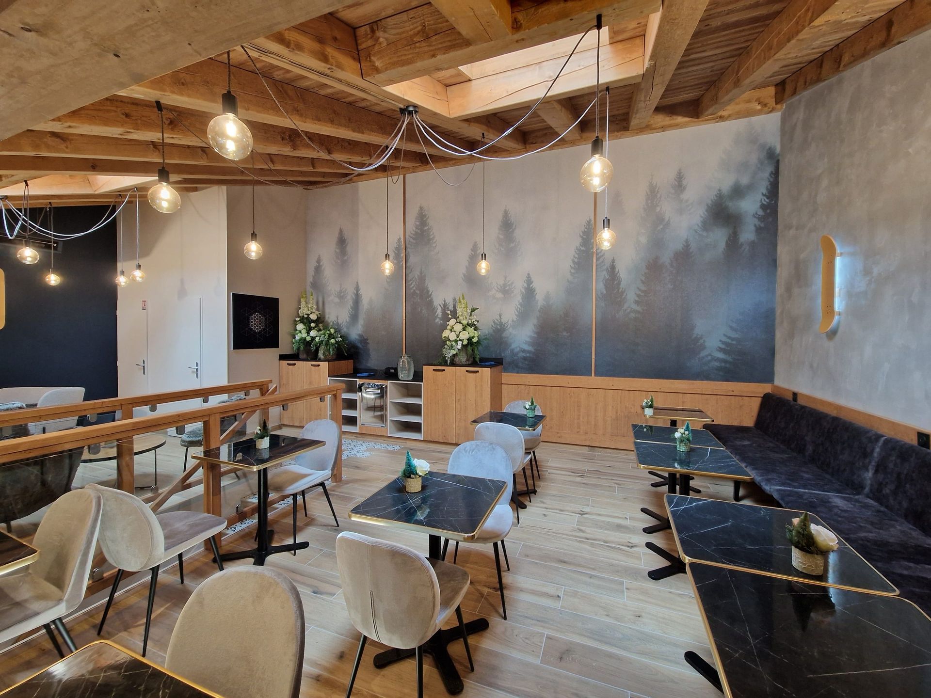 Intérieur du restaurant avec poutres en bois, tables, chaises et une fresque murale représentant une forêt.