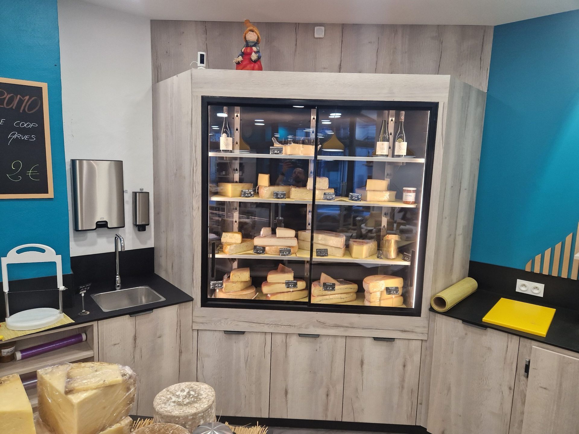 Vitrine à fromages dans un magasin, étagères en bois, fromages variés, murs turquoise et gris clair.