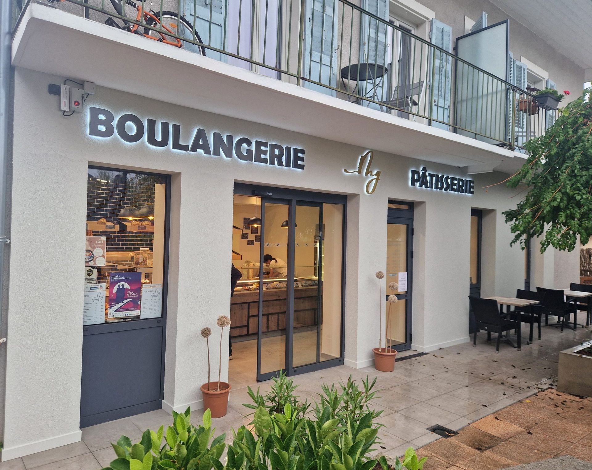 Extérieur d'une boulangerie-pâtisserie avec portes vitrées, murs blancs et terrasse.
