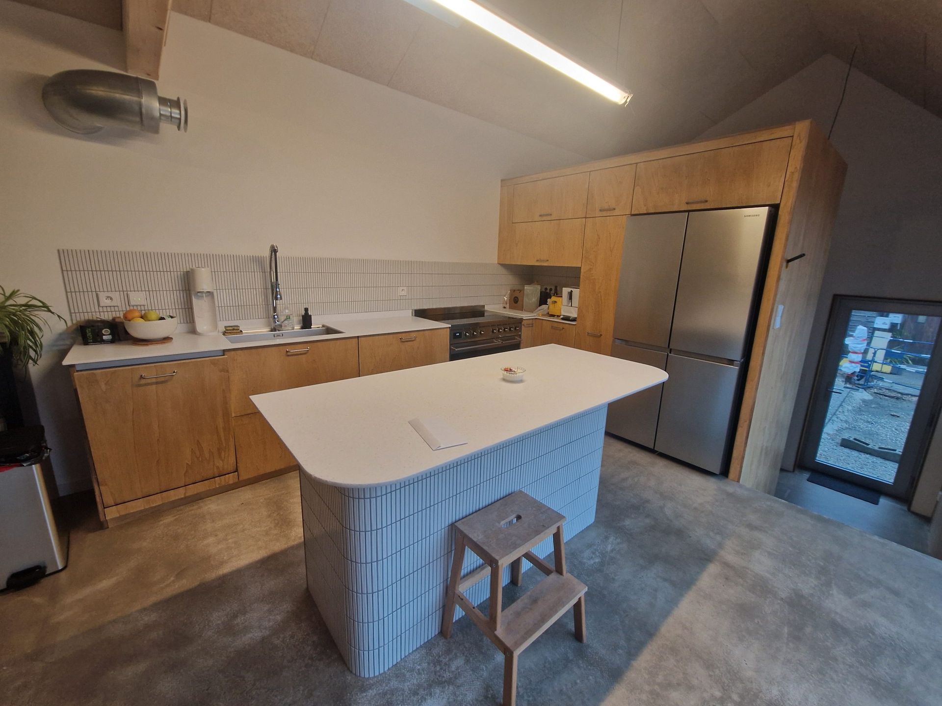 Cuisine moderne avec placards en bois clair, plans de travail blancs et un îlot central avec un petit tabouret en bois.