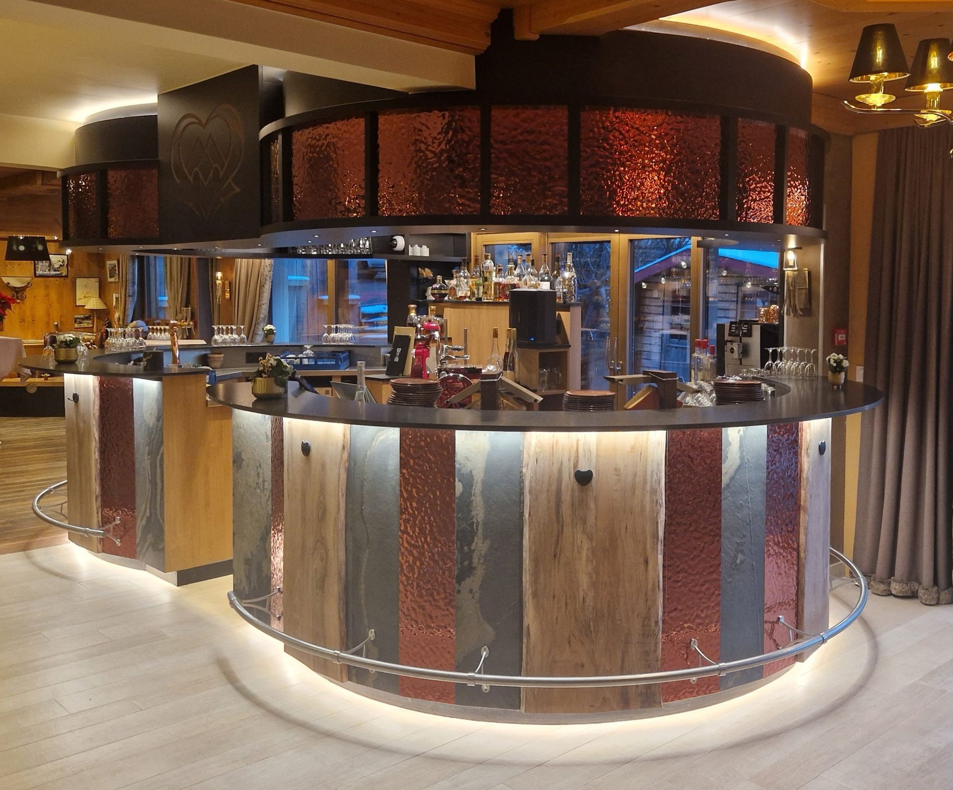 Un bar circulaire à la façade en cuivre, bois et pierre, à l'éclairage tamisé, laissant apparaître le matériel de bar à l'intérieur.