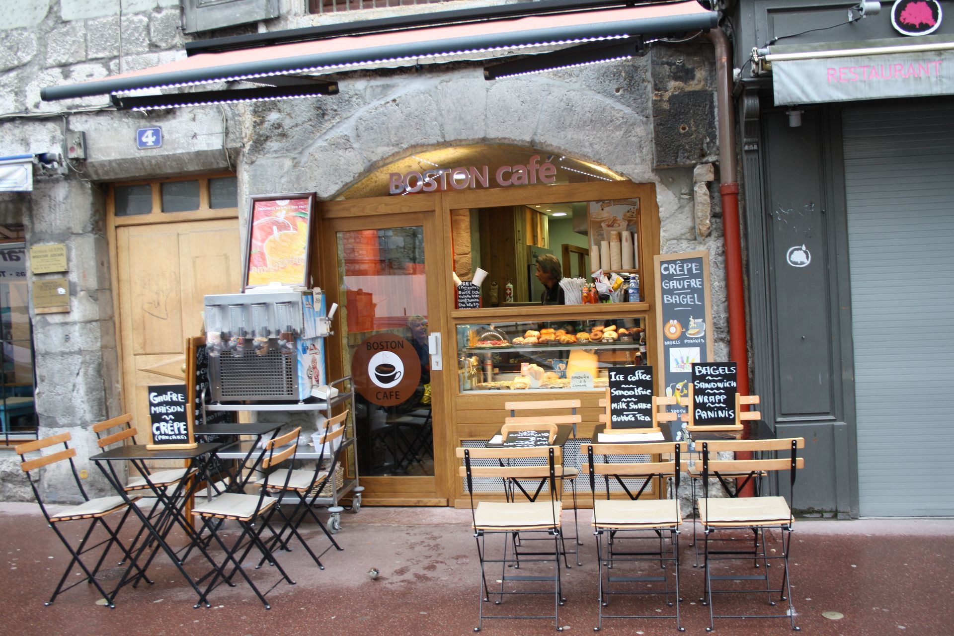 Café avec porte en bois, tables et chaises en terrasse. Enseigne Boston Cafe au-dessus de l'entrée.