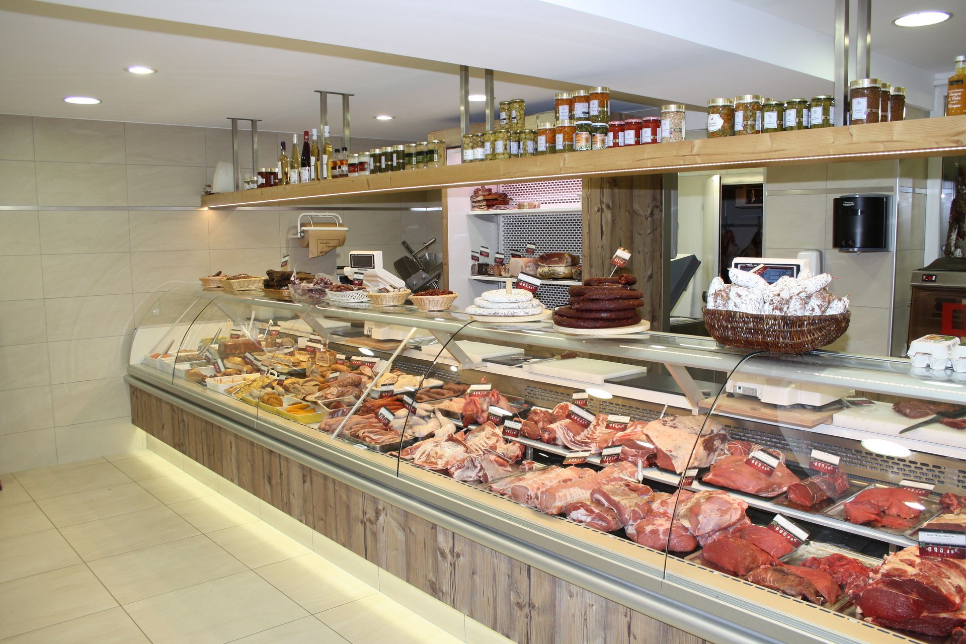 Boucherie avec vitrine présentant diverses viandes et plats préparés. Étagères au-dessus des bocaux et bouteilles exposés.