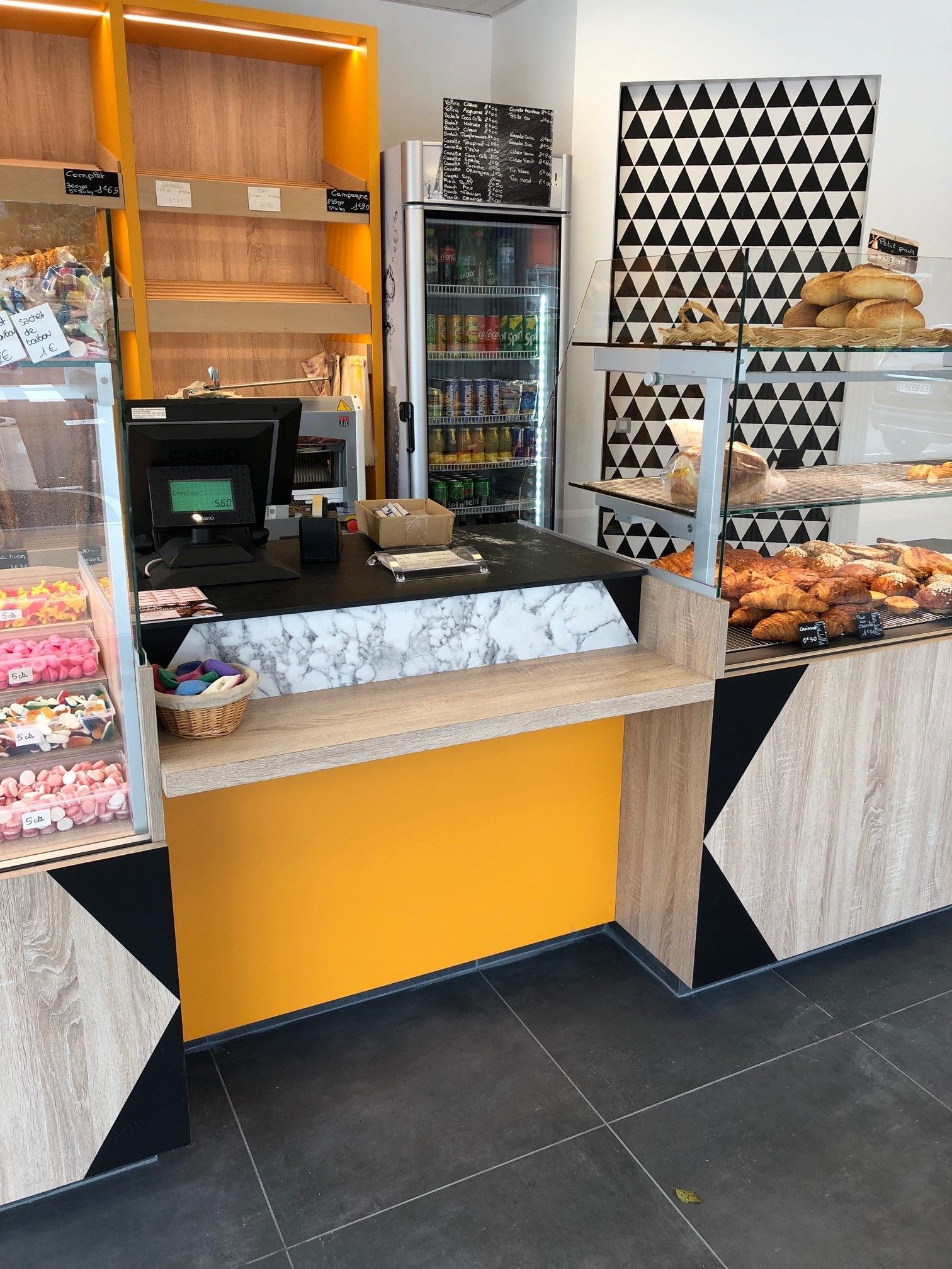 Intérieur de la boulangerie : pâtisseries dans des vitrines, touches de jaune, mur géométrique noir et blanc, boissons dans un réfrigérateur.