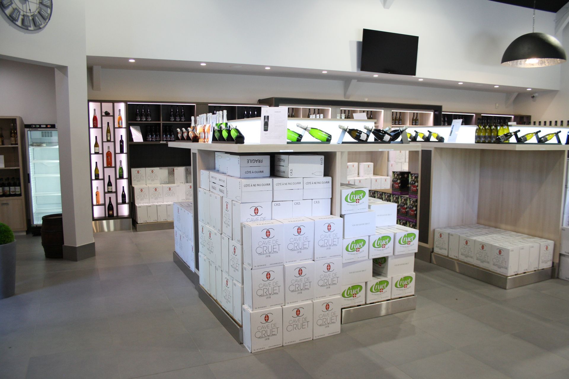 Intérieur d'un magasin de vins. Présentoir de cartons de vin, étagères avec des bouteilles, réfrigérateurs et télévision.