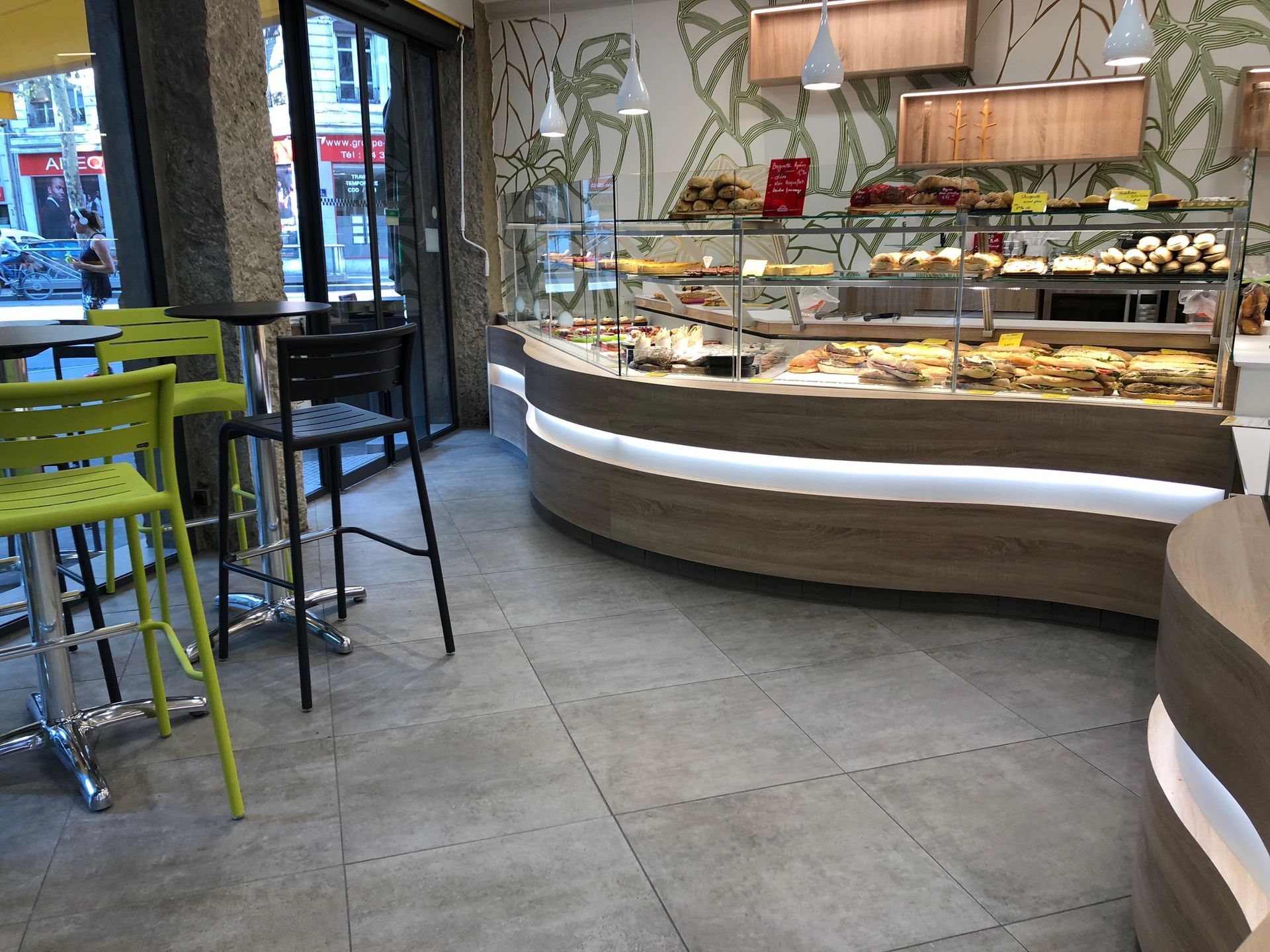 Intérieur d'une boulangerie avec une vitrine de pâtisseries. Tabourets de bar vert citron et noirs, une fenêtre et un mur décoratif.