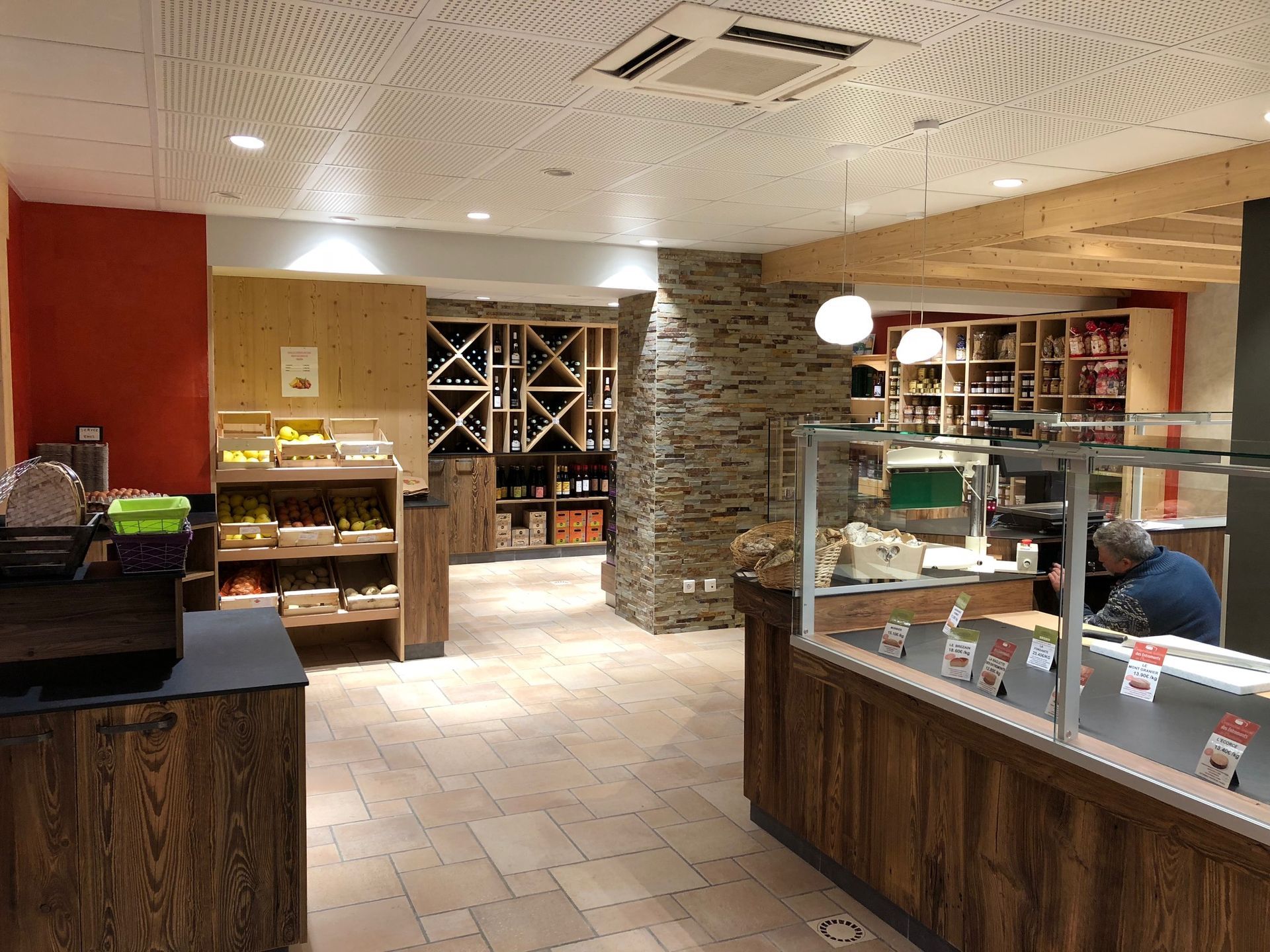 Intérieur d'un magasin, éléments en bois et en pierre, présentoirs de produits alimentaires, casiers à vin, client au comptoir.