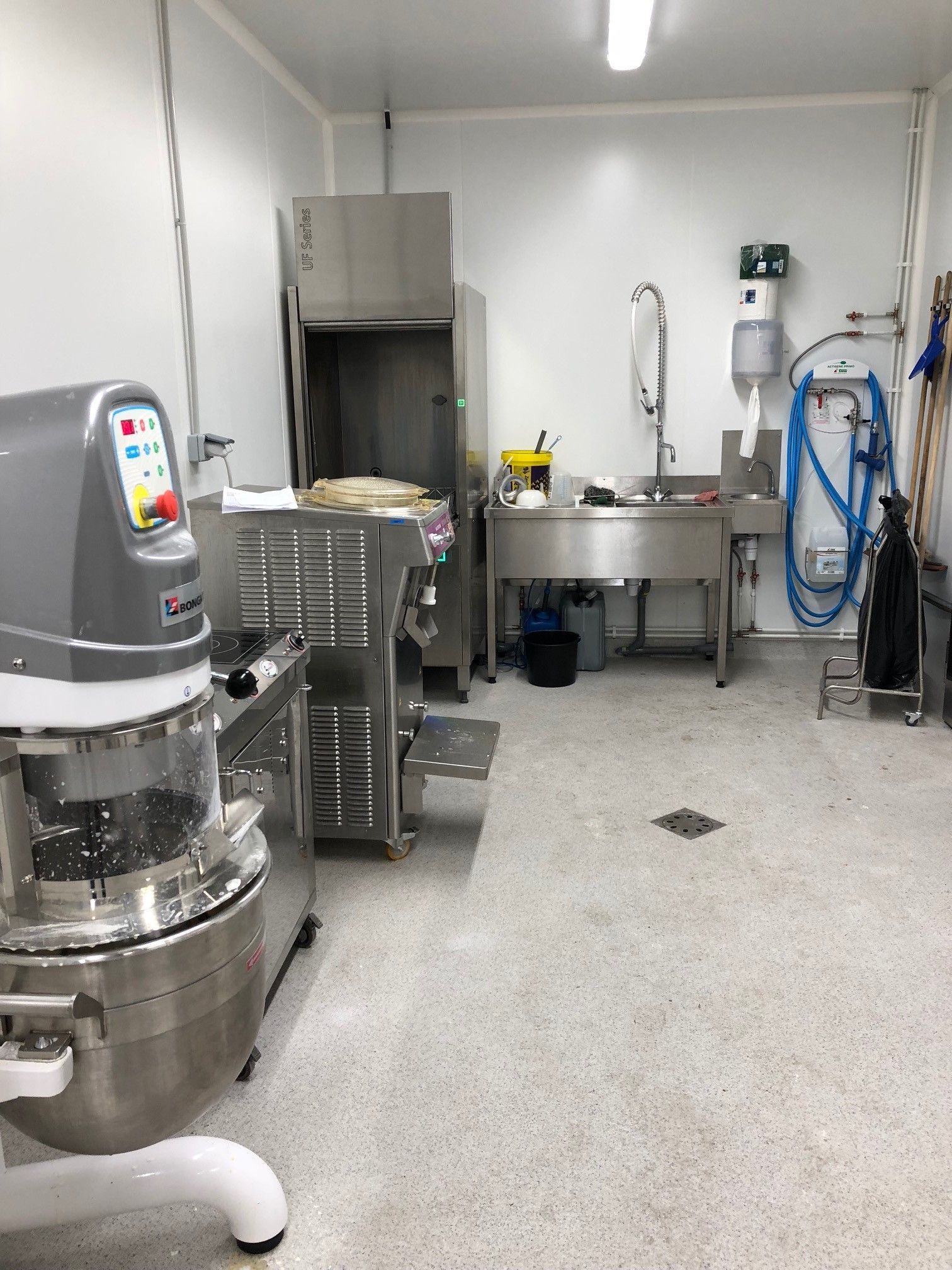 Cuisine professionnelle équipée de matériel en acier inoxydable : mixeur, lave-vaisselle, évier et tuyau d’arrosage, le tout adossé à un mur blanc.