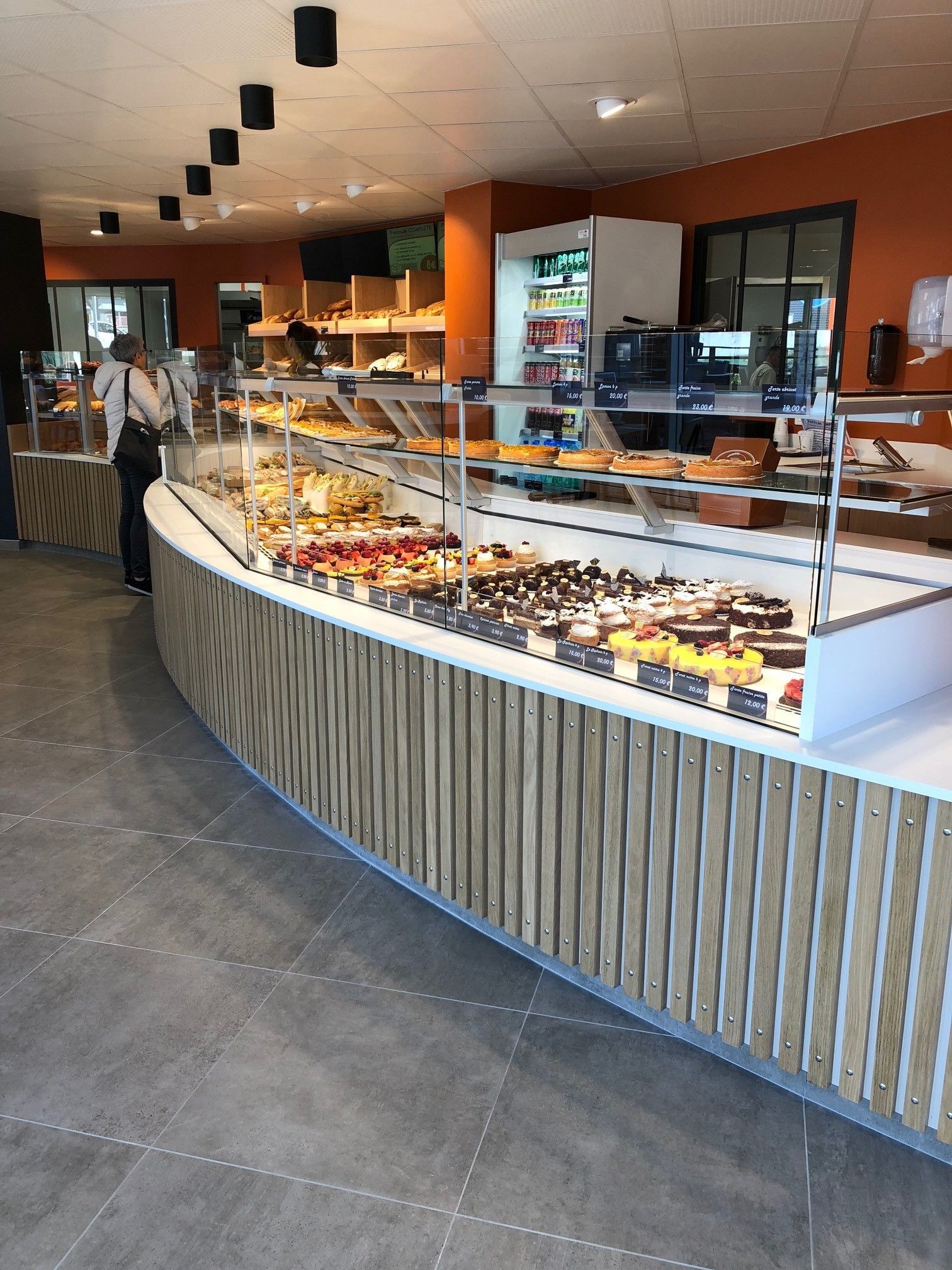 Intérieur d'une boulangerie avec une vitrine incurvée remplie de pâtisseries. Une personne est visible près de la vitrine.