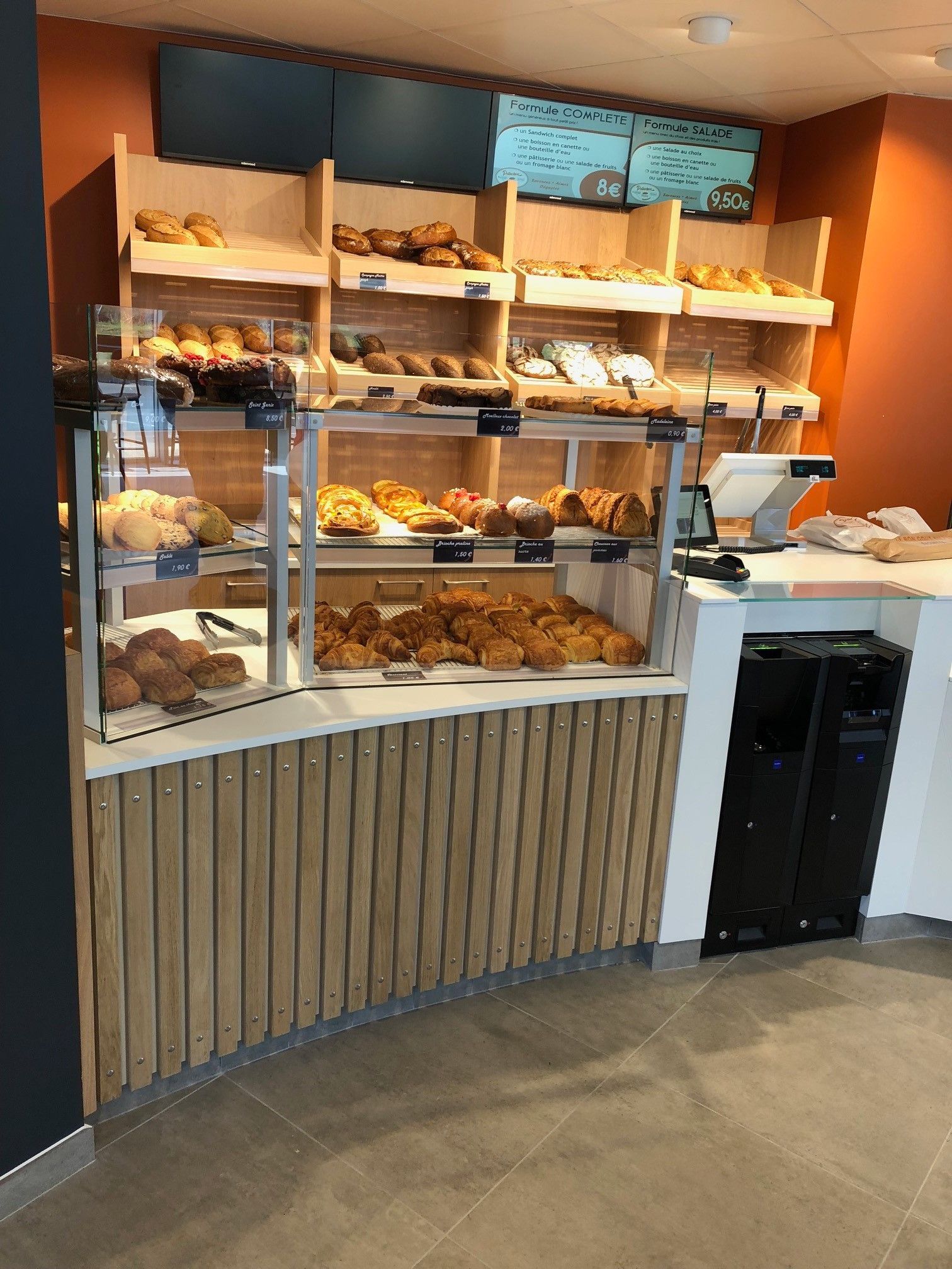 Vitrine de boulangerie remplie de pâtisseries, étagères en bois présentant des produits de boulangerie. Touches d'orange et de bois clair.