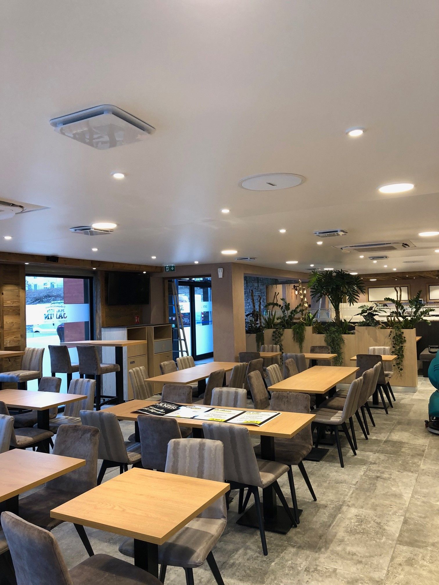 Intérieur de restaurant avec tables et chaises, palette de couleurs neutres, bien éclairé, quelques plantes visibles.