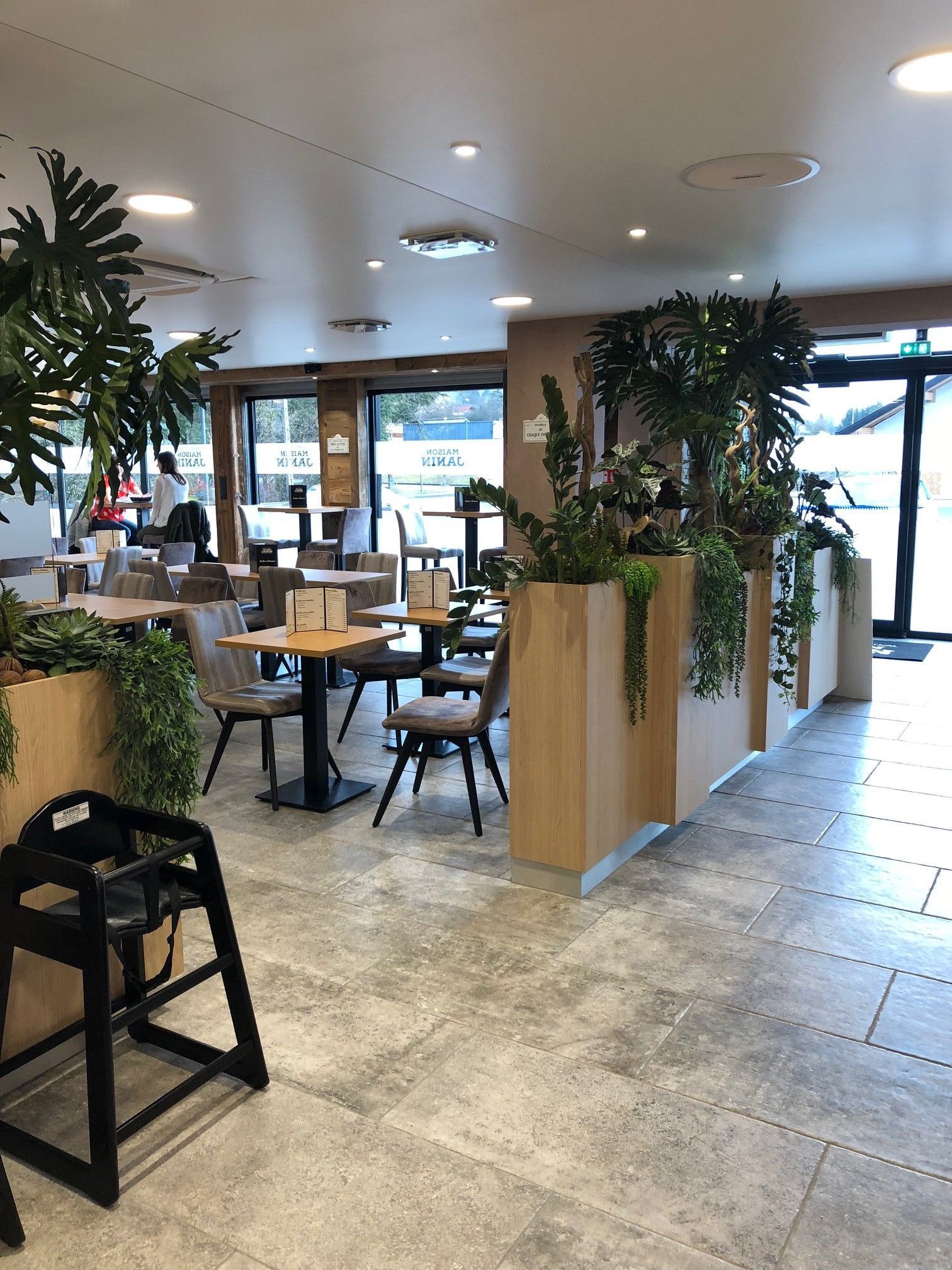 Intérieur d'un café avec tables, chaises et plantes. Sol en bois clair et en pierre grise. Chaise haute pour enfant au premier plan.