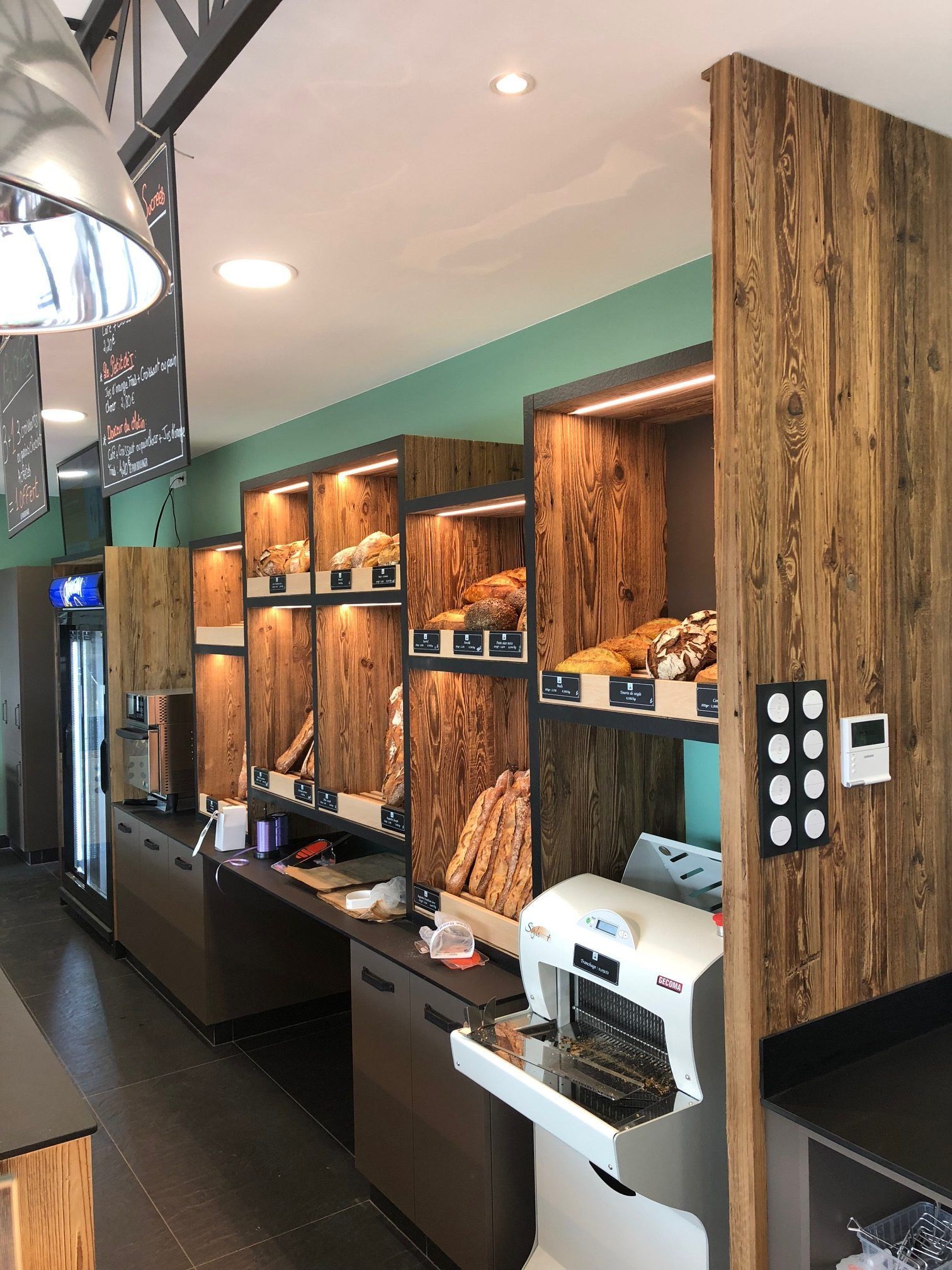 Intérieur de la boulangerie avec des étagères en bois présentant du pain, une trancheuse à pain et une chambre froide.