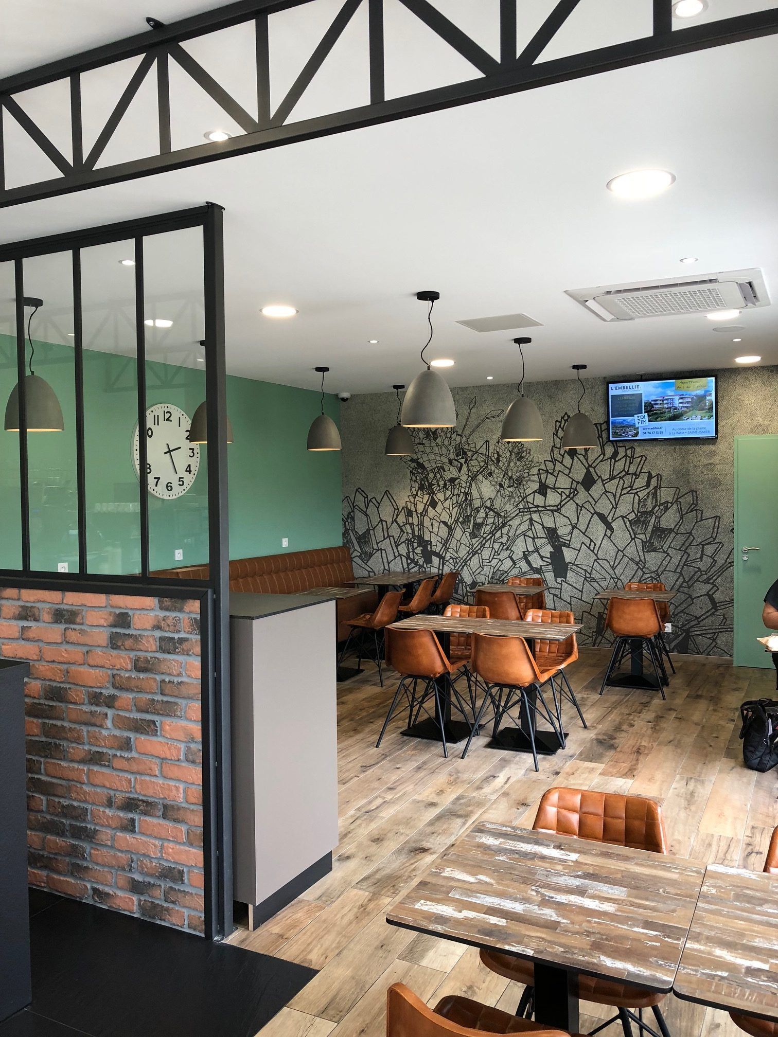 Intérieur du restaurant : tables, chaises, suspensions lumineuses, murs en briques et verts, mur d’accent à motifs et parquet.