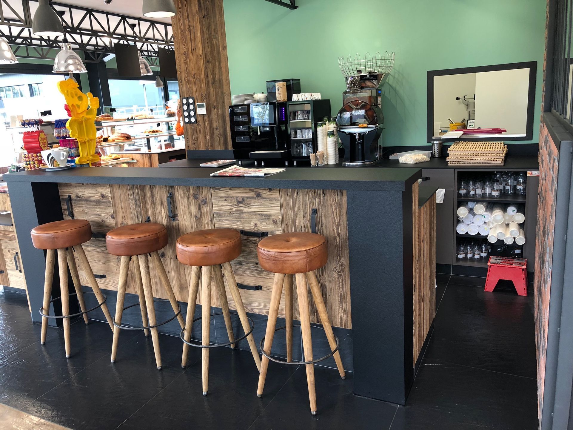 Coin café avec des accents de bois et de noir, quatre tabourets, une machine à café, un miroir et quelques produits.