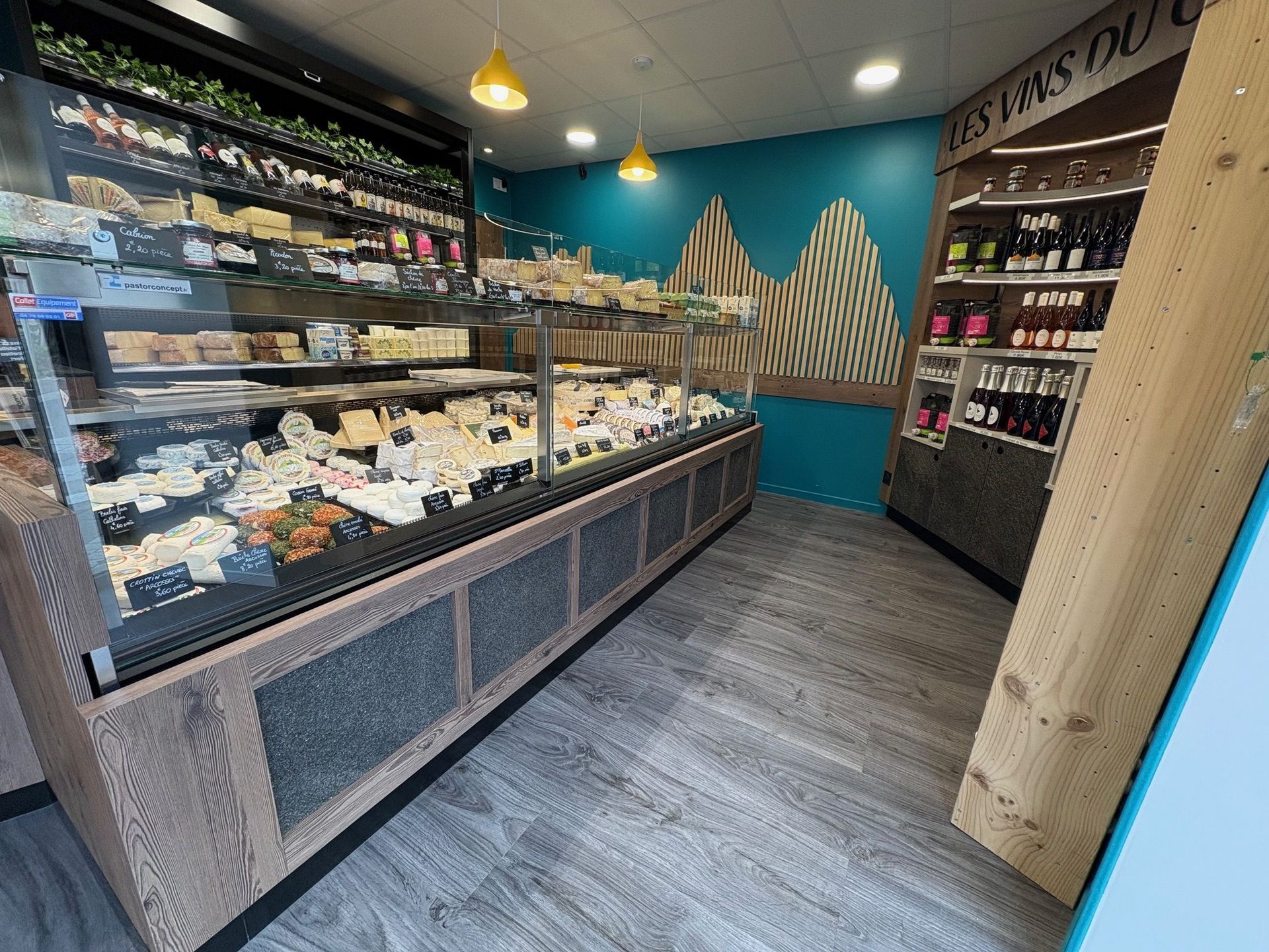 Intérieur d'une fromagerie avec une vitrine remplie de fromages, un mur turquoise et des éléments en bois.