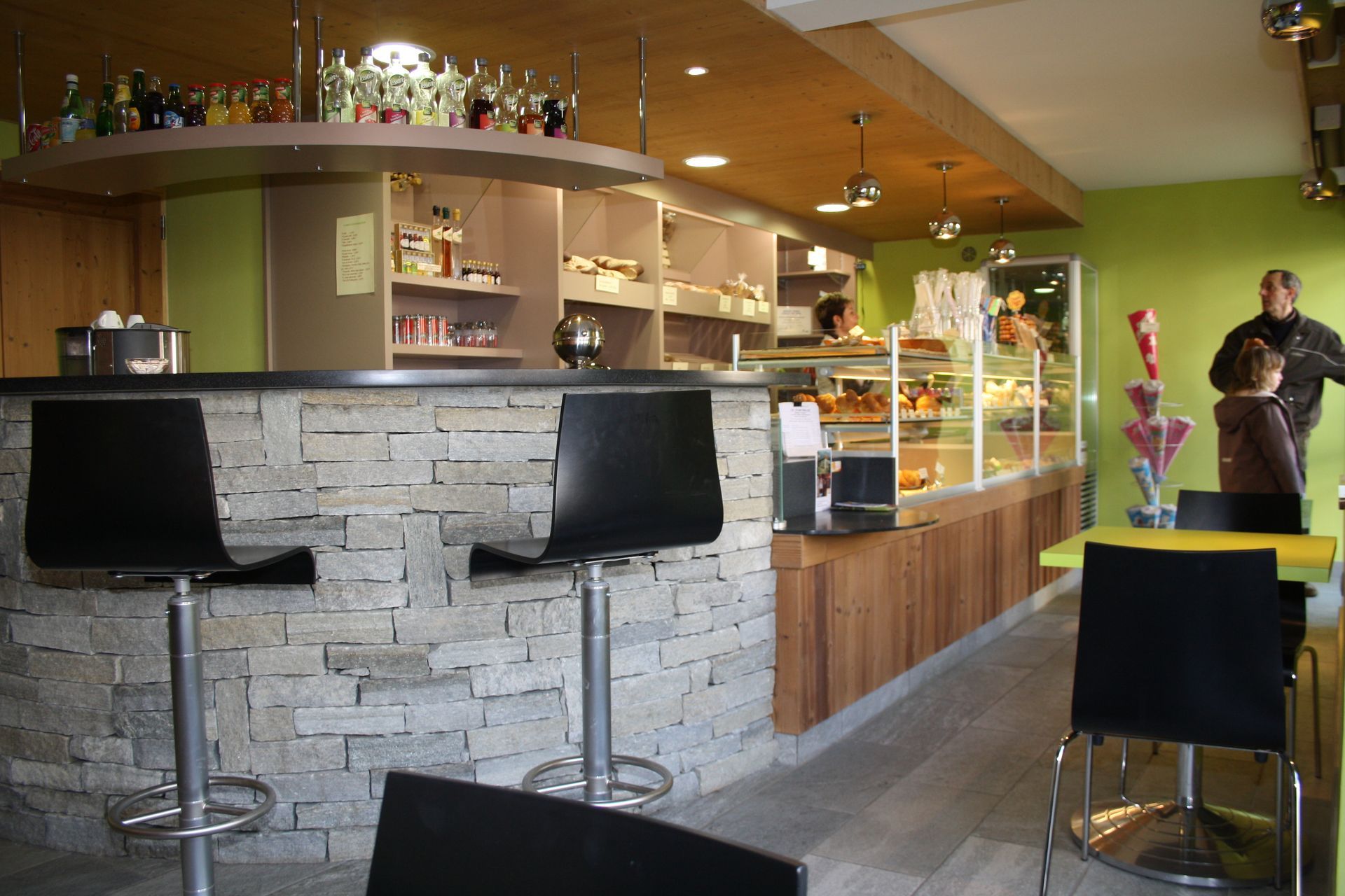 Intérieur d'un café avec un bar en pierre, des tabourets et une vitrine de pâtisseries ; des clients se tiennent près d'une table.