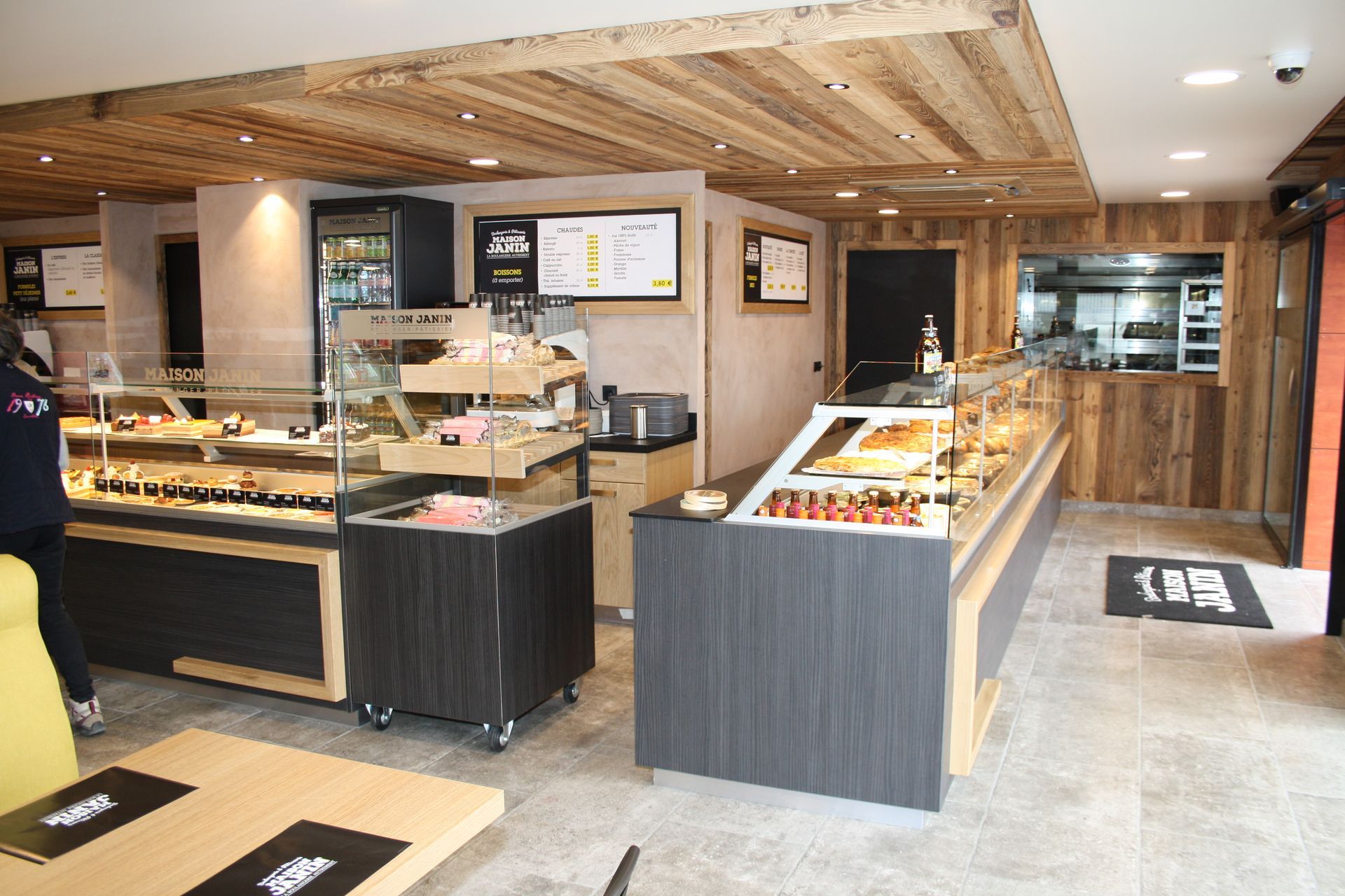Intérieur d'une boulangerie avec vitrines à pâtisseries, plafond en bois et cuisine visible par une fenêtre.