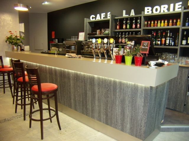 Bar du « Café La Borie » avec tabourets. Façade grise avec bandeau lumineux, mur du fond noir et étagères à bouteilles.