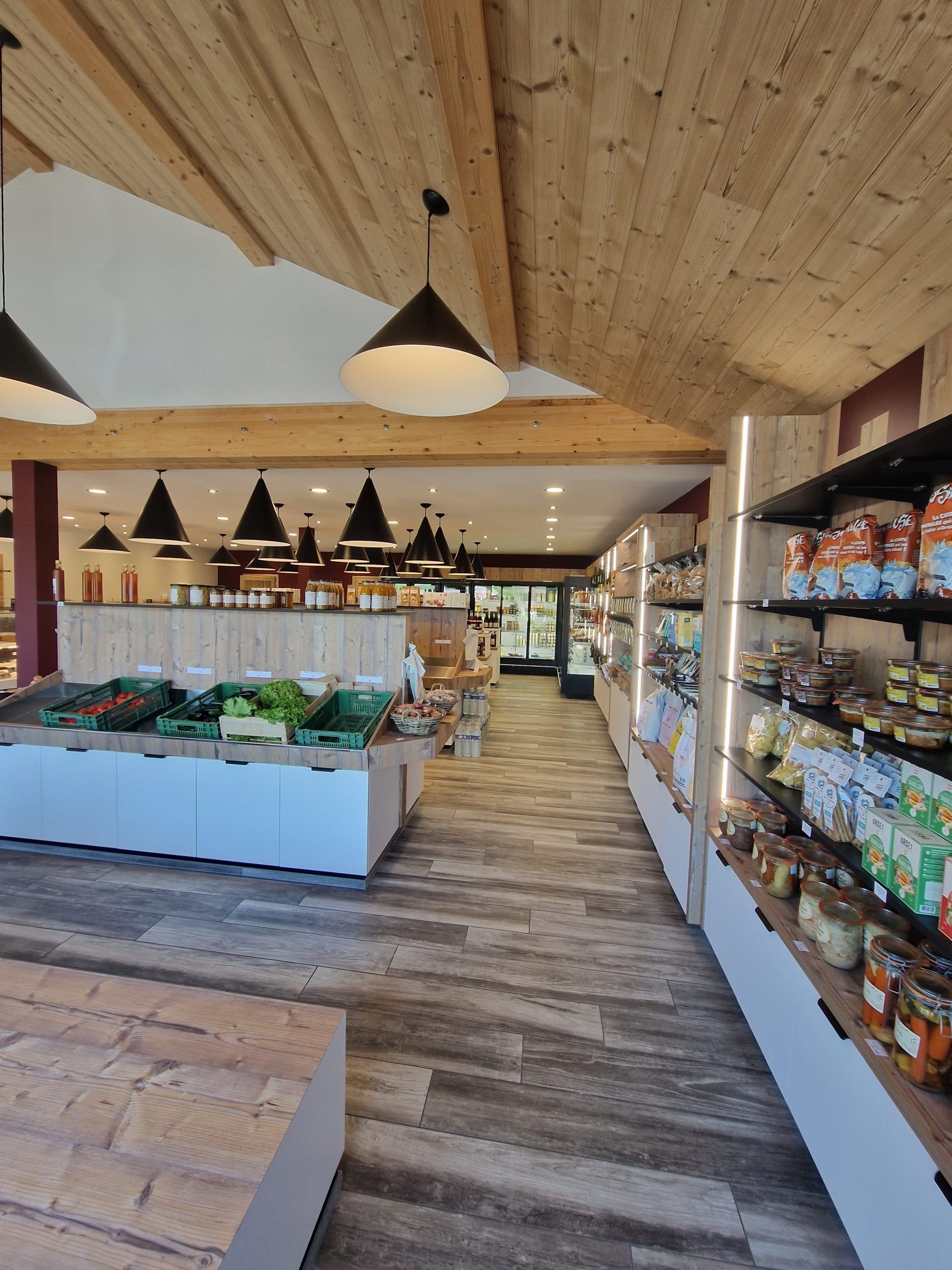 Intérieur d'un magasin d'alimentation avec étalage de fruits et légumes et étagères de produits emballés, design en bois et lumière.
