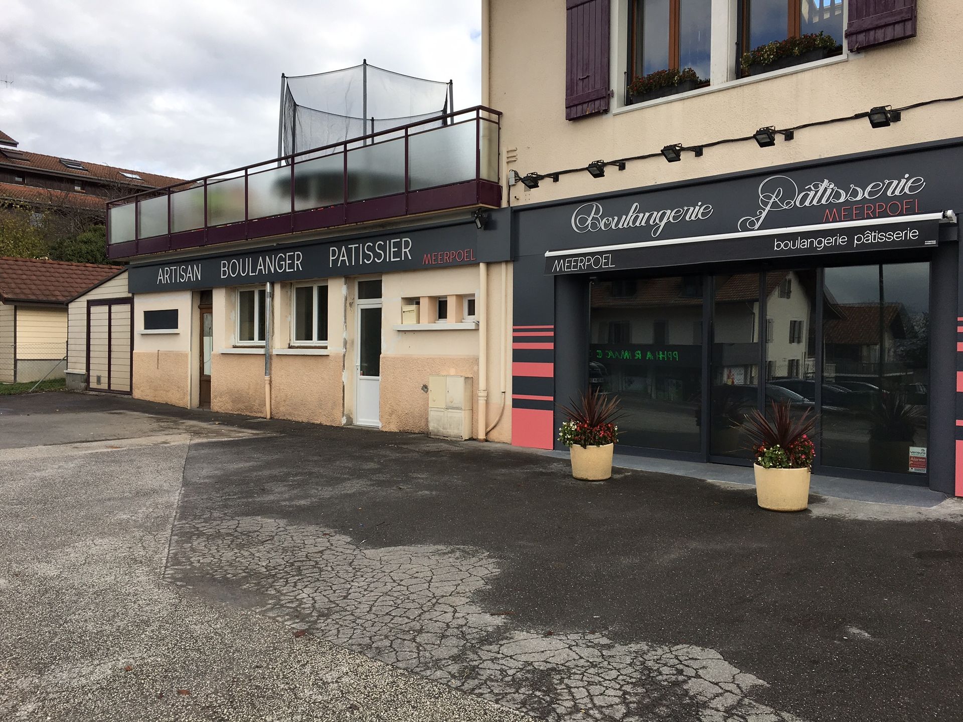 Boulangerie à la façade grise et rose, balcon, plantes en pot. Enseigne « Boulangerie Pâtisserie » au-dessus de la porte.