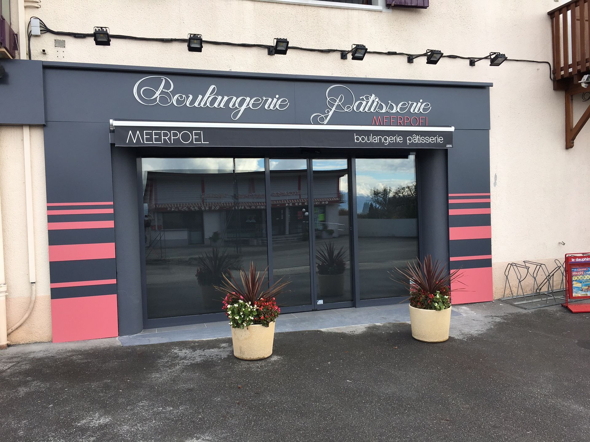 Entrée de boulangerie avec façade grise, auvent et portes vitrées. Détails rayés rouges et noirs, deux plantes en pot.