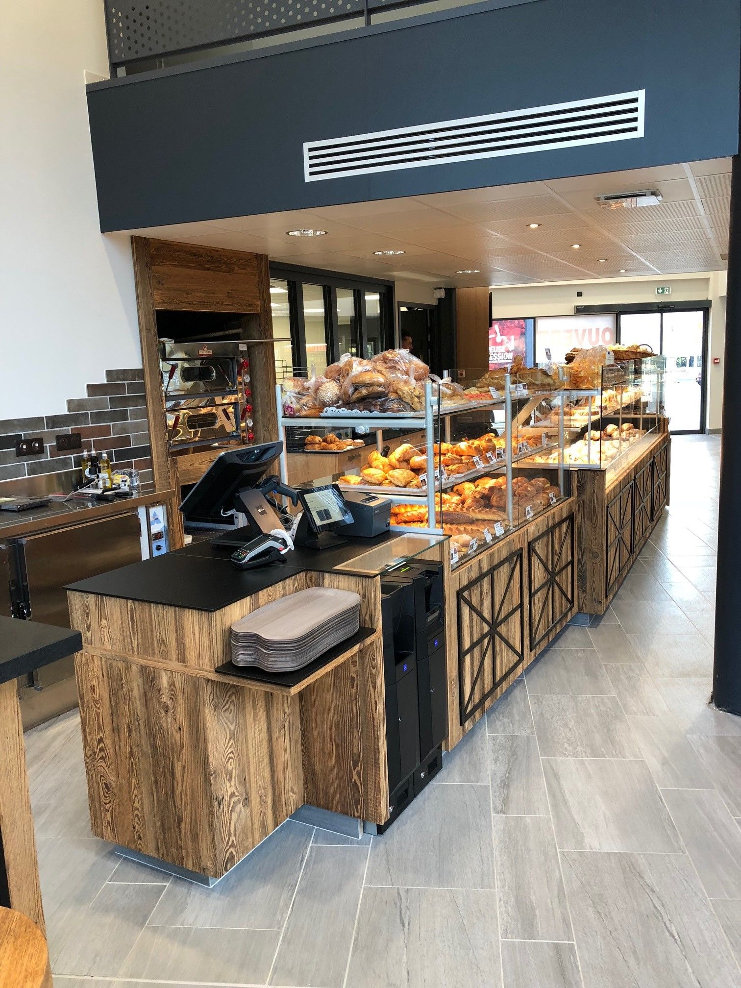 Vitrine de boulangerie présentant divers produits de boulangerie. Comptoir en bois avec caisse enregistreuse. Intérieur lumineux.