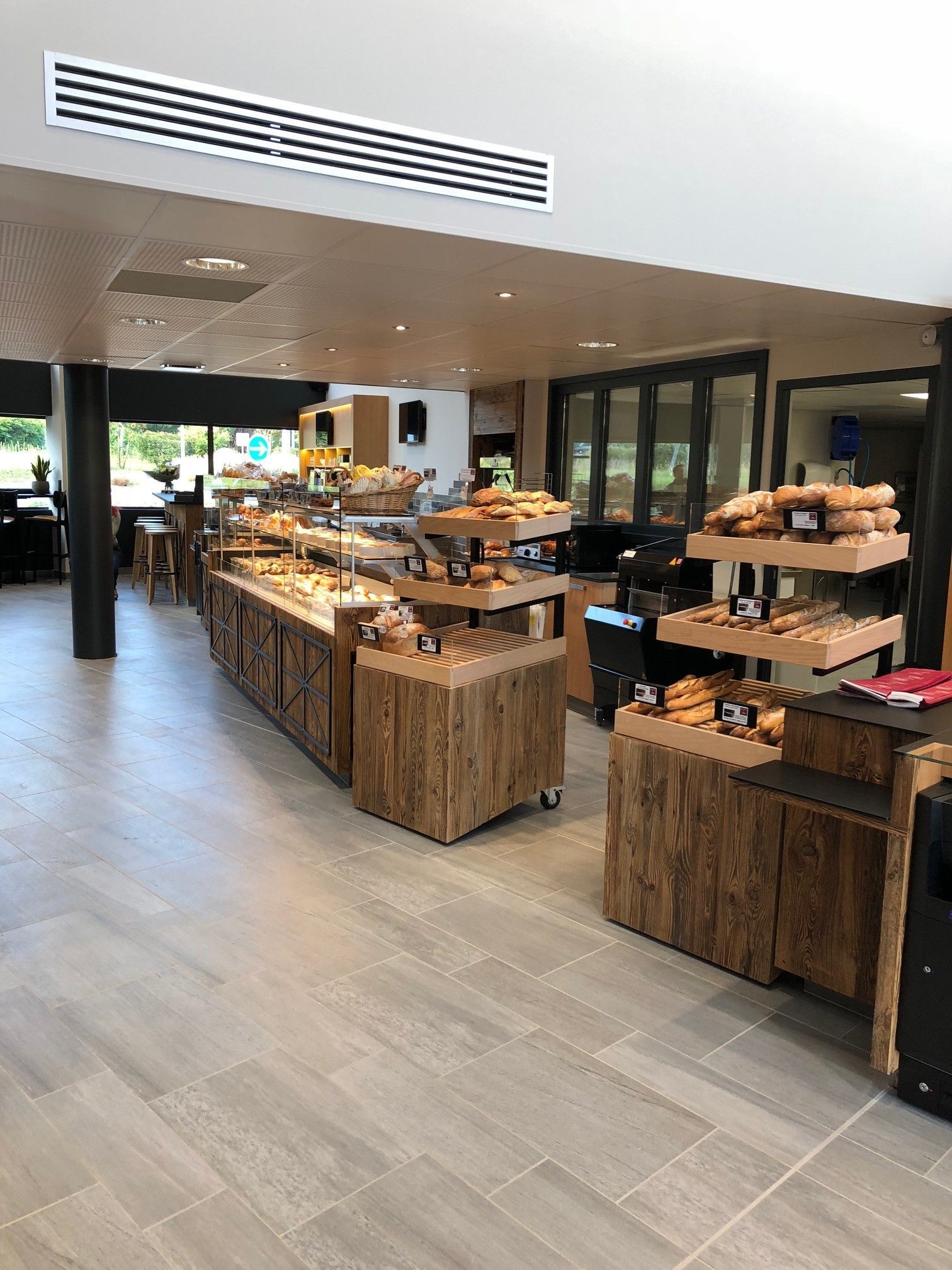 Intérieur d'une boulangerie avec vitrines en bois présentant pains et viennoiseries. Sol carrelé gris, éclairage encastré et vue sur l'extérieur.