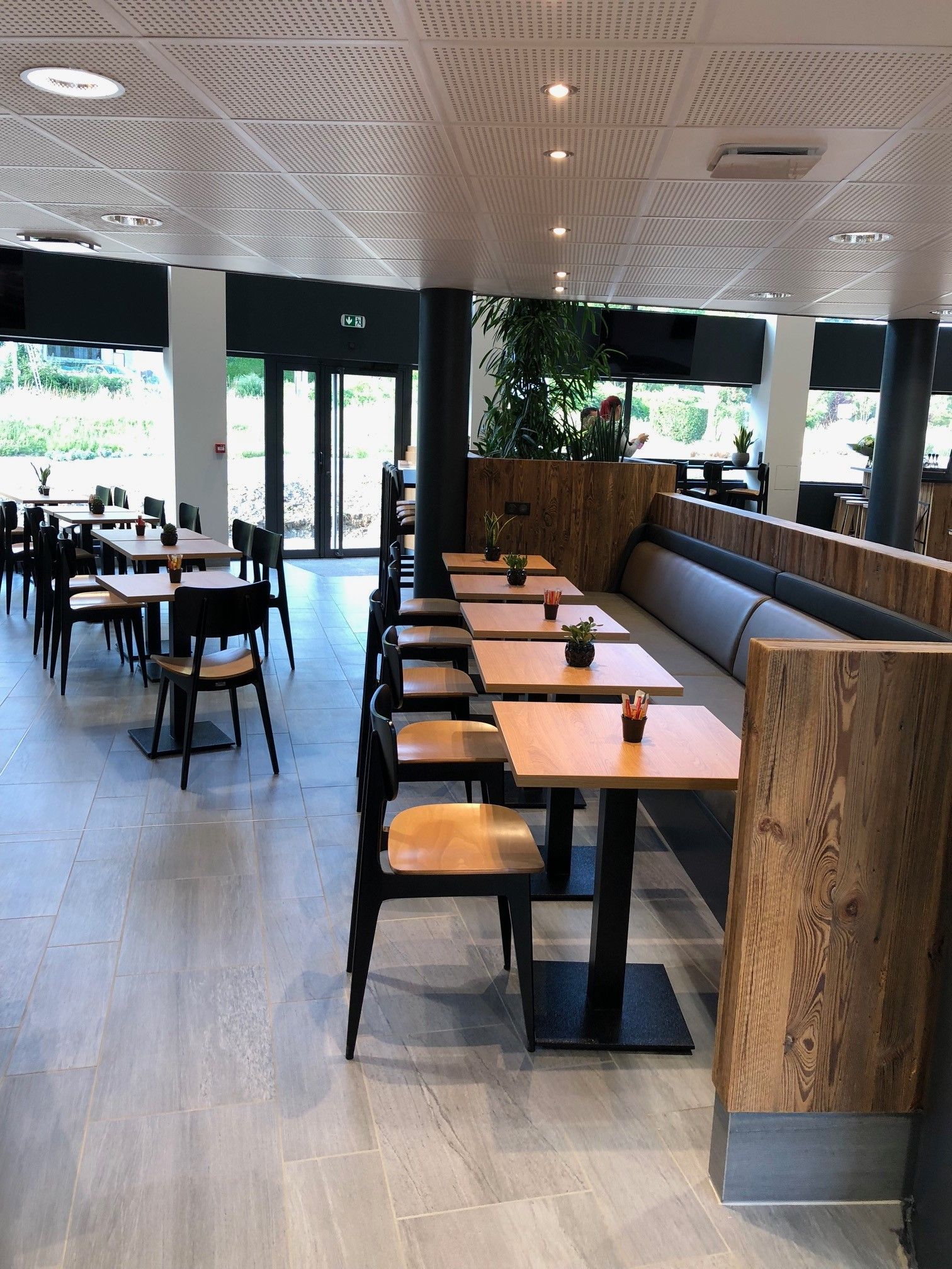 Intérieur d'un restaurant vide, avec des tables, des chaises et une longue banquette intégrée. La lumière naturelle y pénètre abondamment.