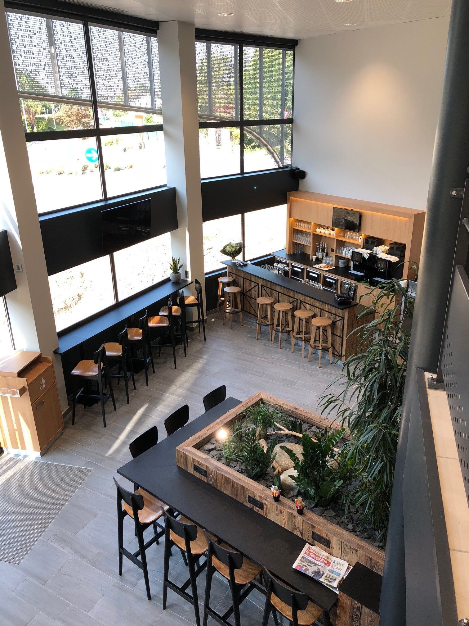 Vue en plongée de l'intérieur d'un café moderne avec un bar, des sièges et une grande composition végétale. De grandes fenêtres laissent entrer la lumière naturelle.