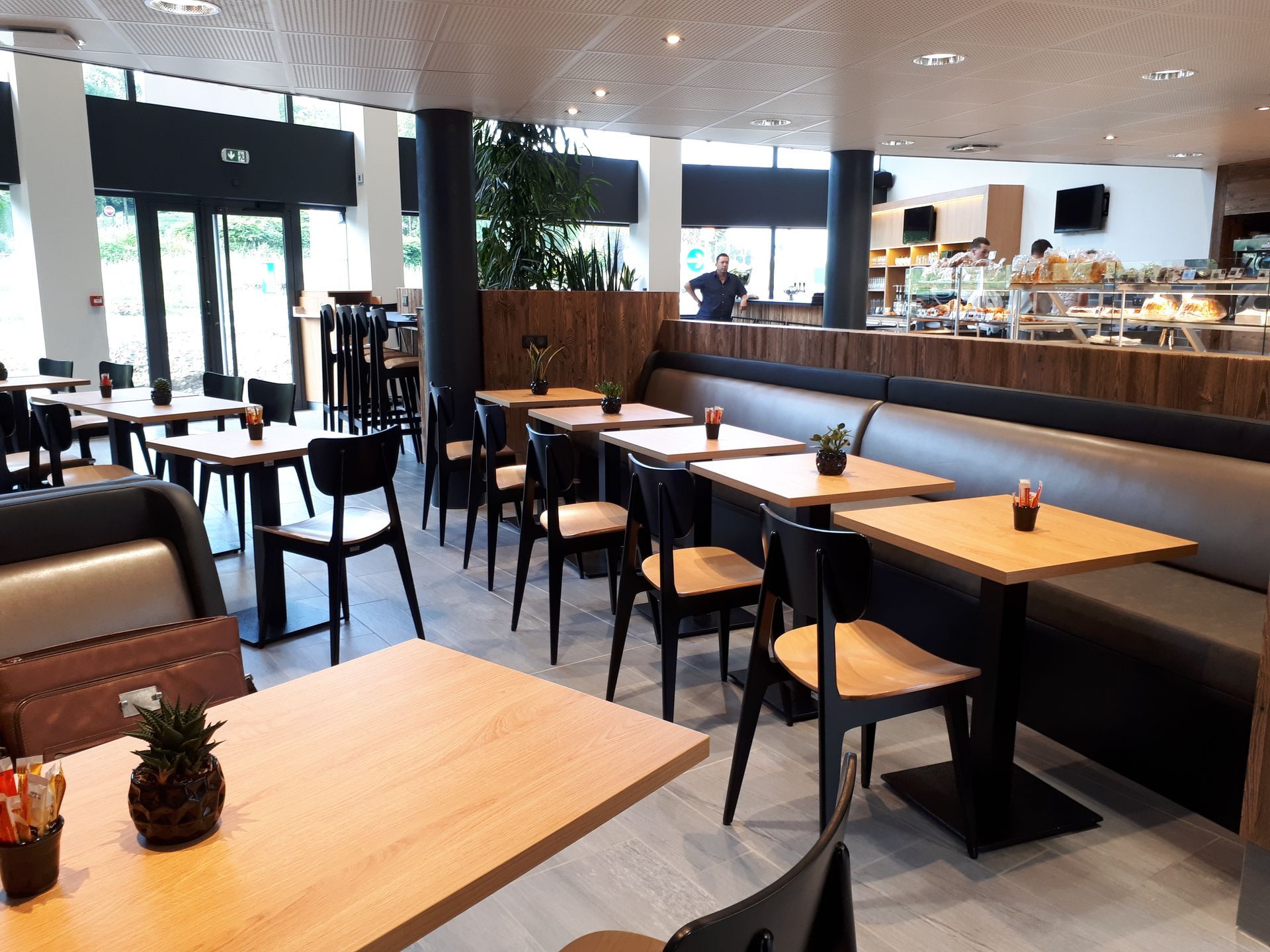 Intérieur d'un café avec tables, chaises et une longue banquette. On aperçoit des éléments en bois et un comptoir où sont présentés des plats.