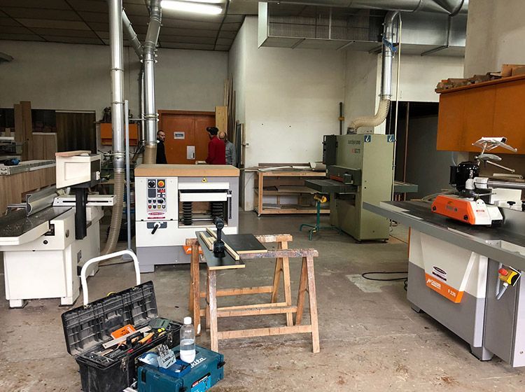 Atelier avec machines à bois, une personne en chemise rouge, des outils et une planche de bois sur un tréteau.