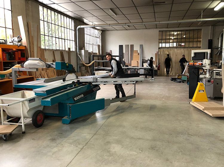 Un atelier de menuiserie avec un ouvrier manipulant une grande scie. Plusieurs autres personnes et du matériel sont visibles.